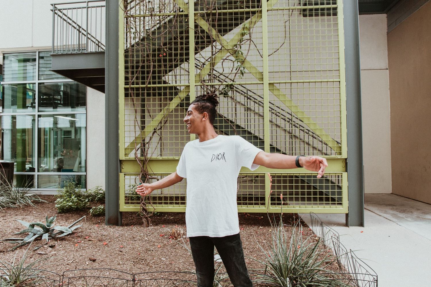 Man in white t-shirt posing with arms outstretched outdoors near modern building with yellow metal staircase.