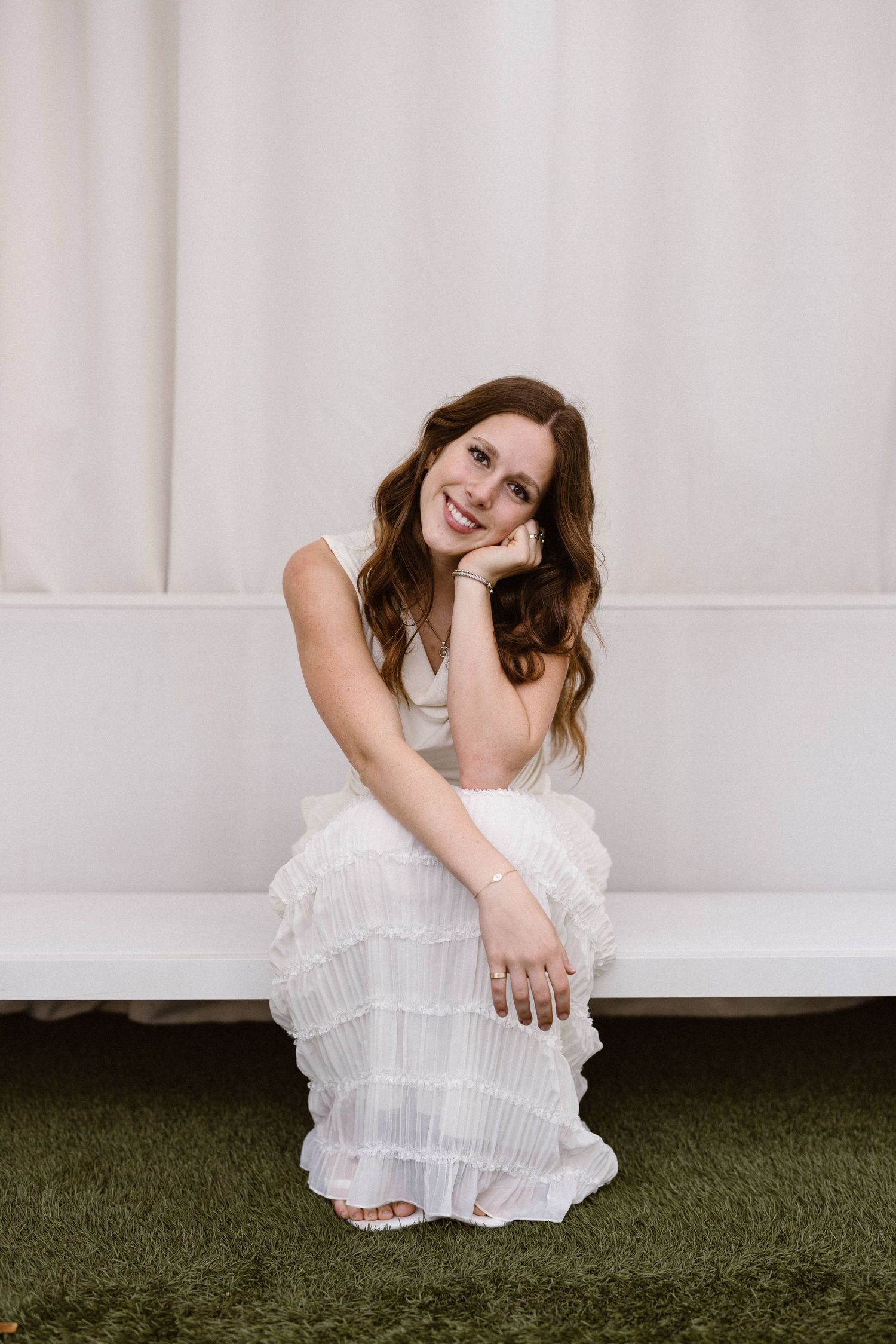 Smiling young woman in a white ruffled dress crouching on green grass in front of a white curtain backdrop.