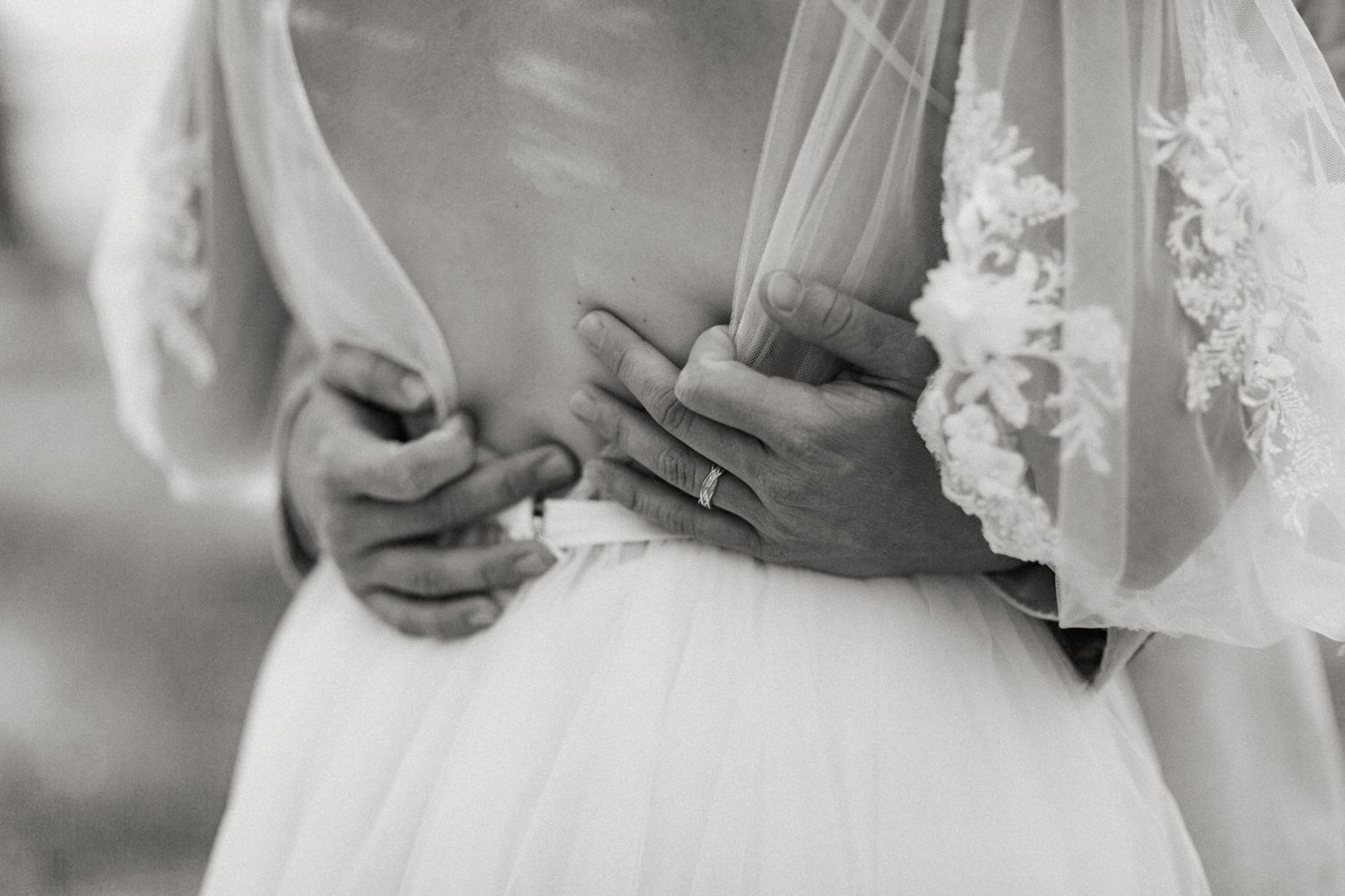 Black and white close-up of bride and groom hands embracing at waist, wedding ring visible on lace gown.