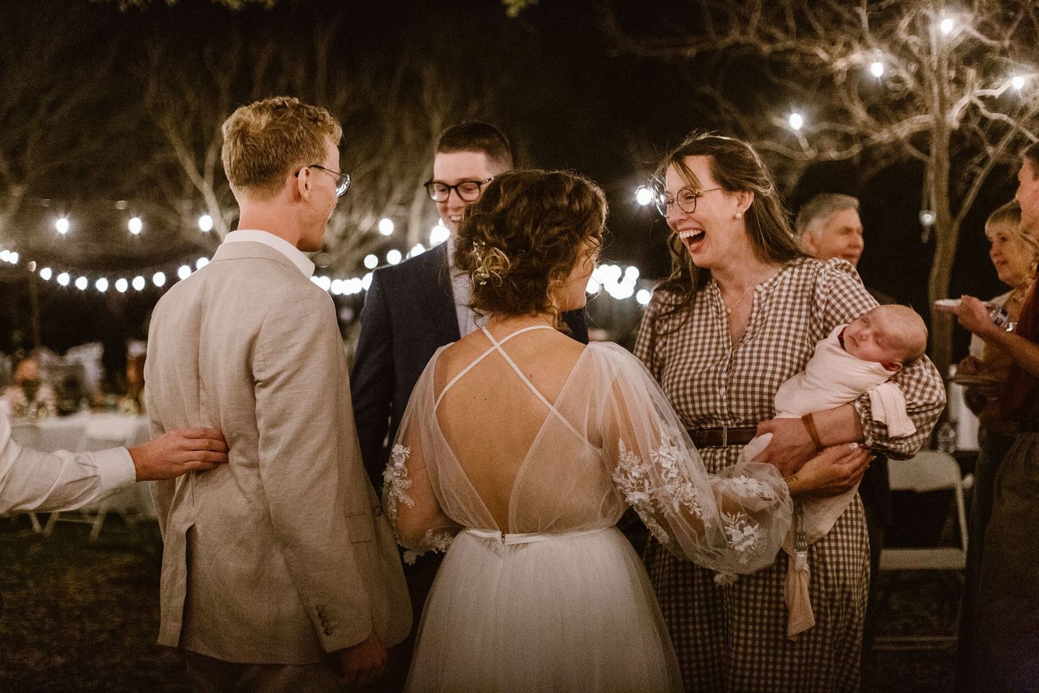 Bride and groom greeting guests at outdoor evening wedding reception under string lights.