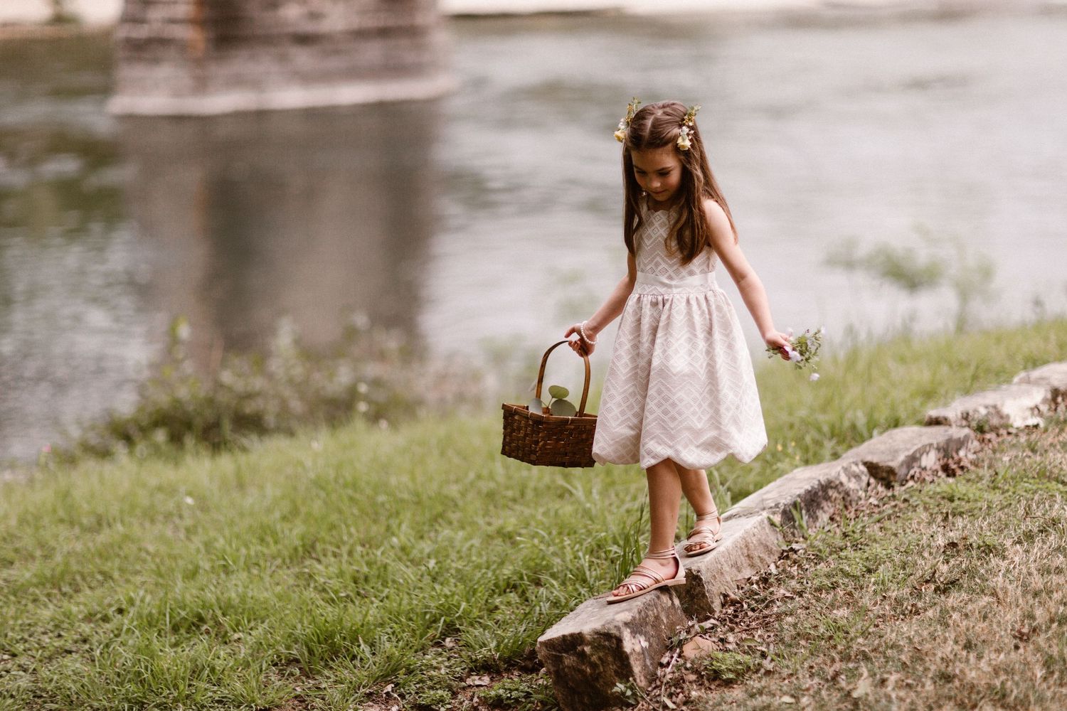 A young girl in a white dress and floral crown carries a basket near a river.