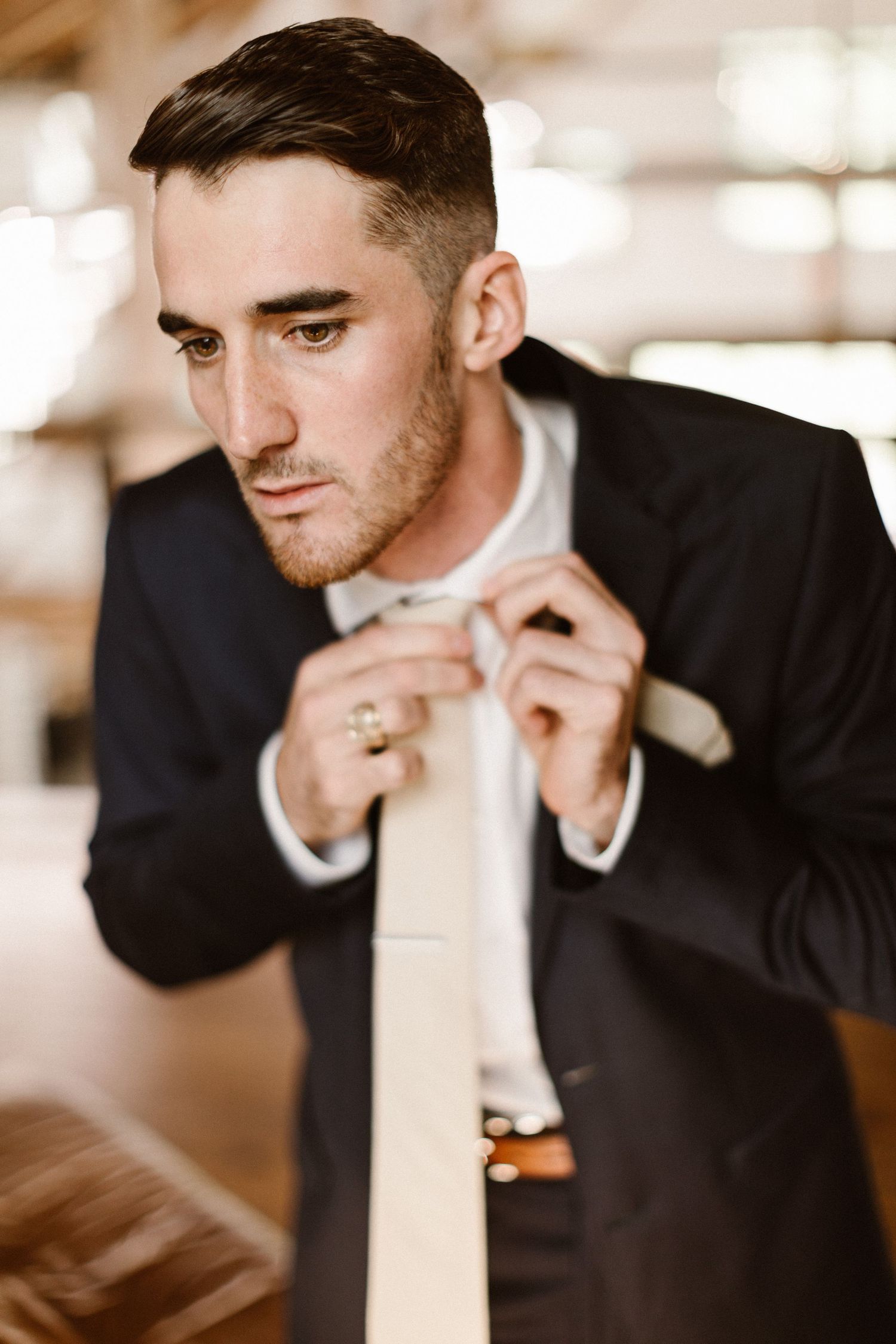 Dapper groom in navy suit adjusting his cream tie while getting ready indoors.
