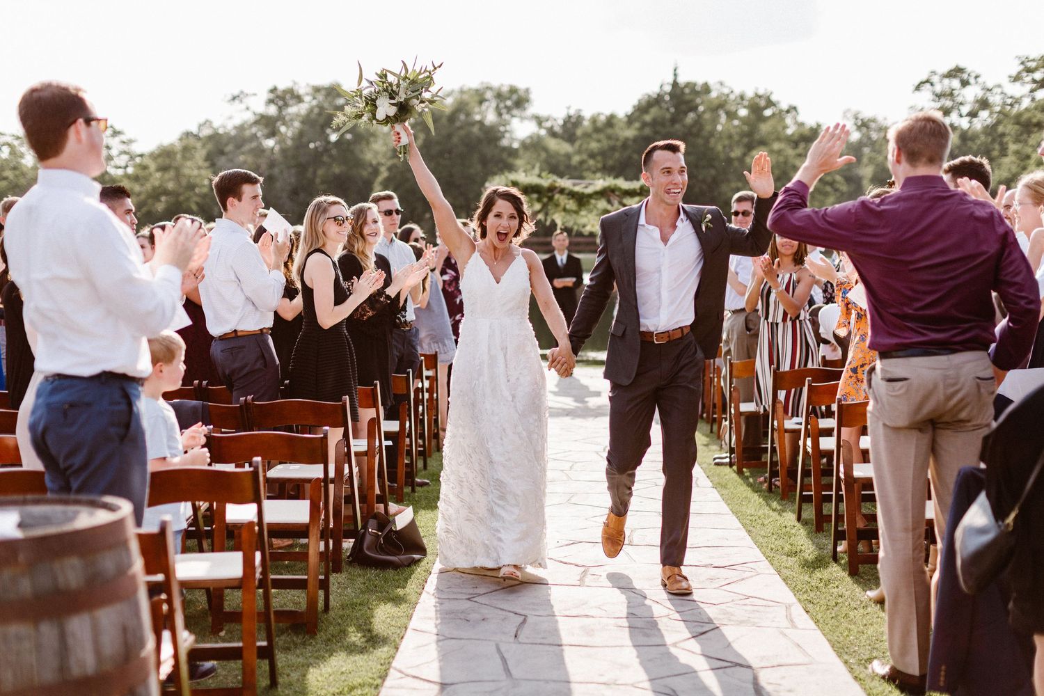 Joyful newlyweds walk back down aisle as guests applaud at sunny outdoor wedding ceremony.