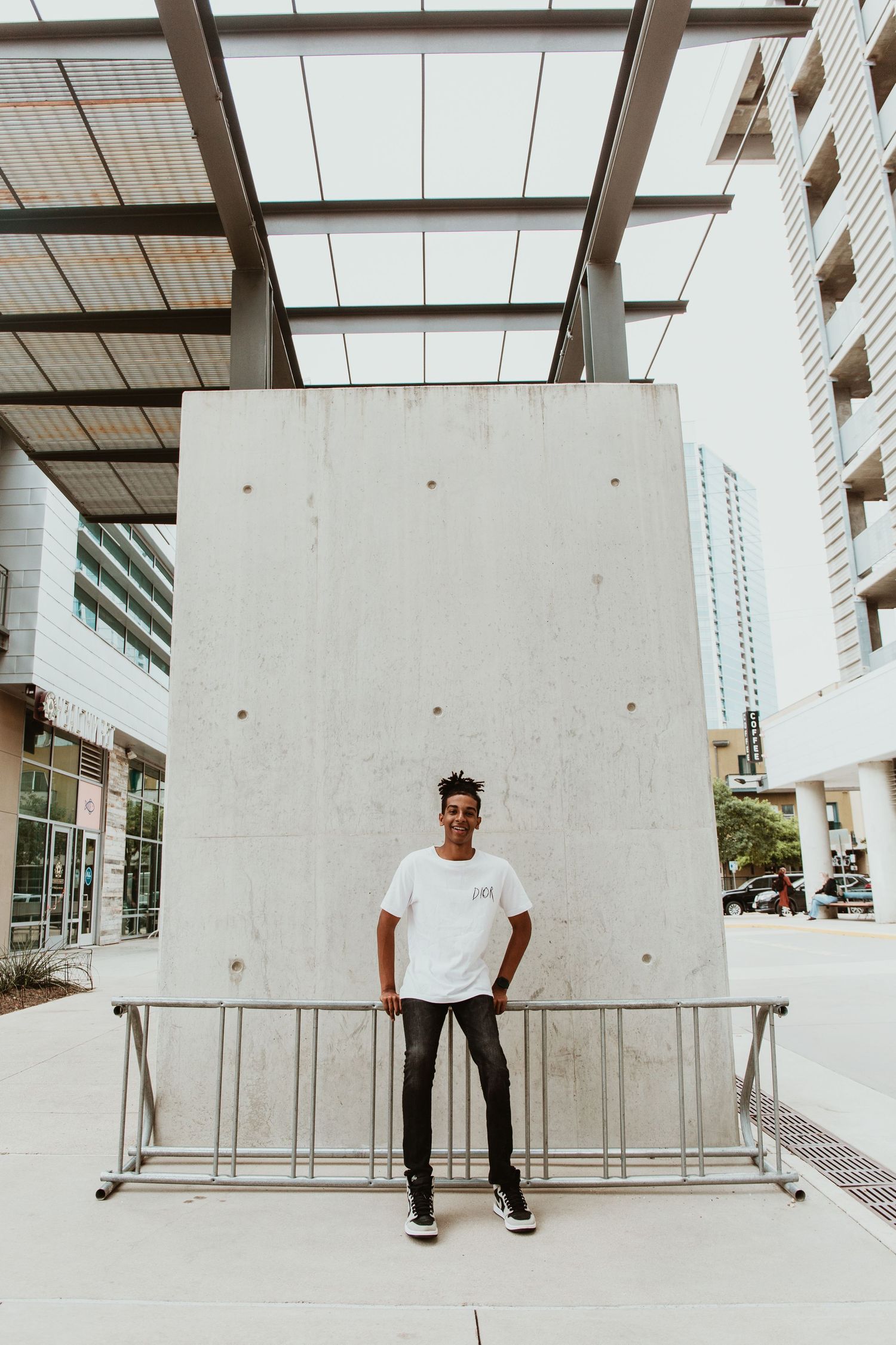 Young man in white t-shirt and black jeans standing confidently in front of a concrete wall in an urban setting.