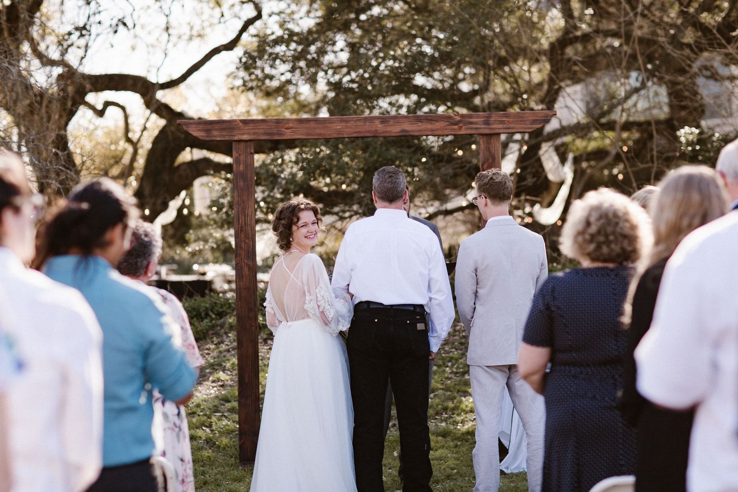 Bride in a lace-back gown and groom exchange vows under a wooden arch with oak trees and string lights in the background.