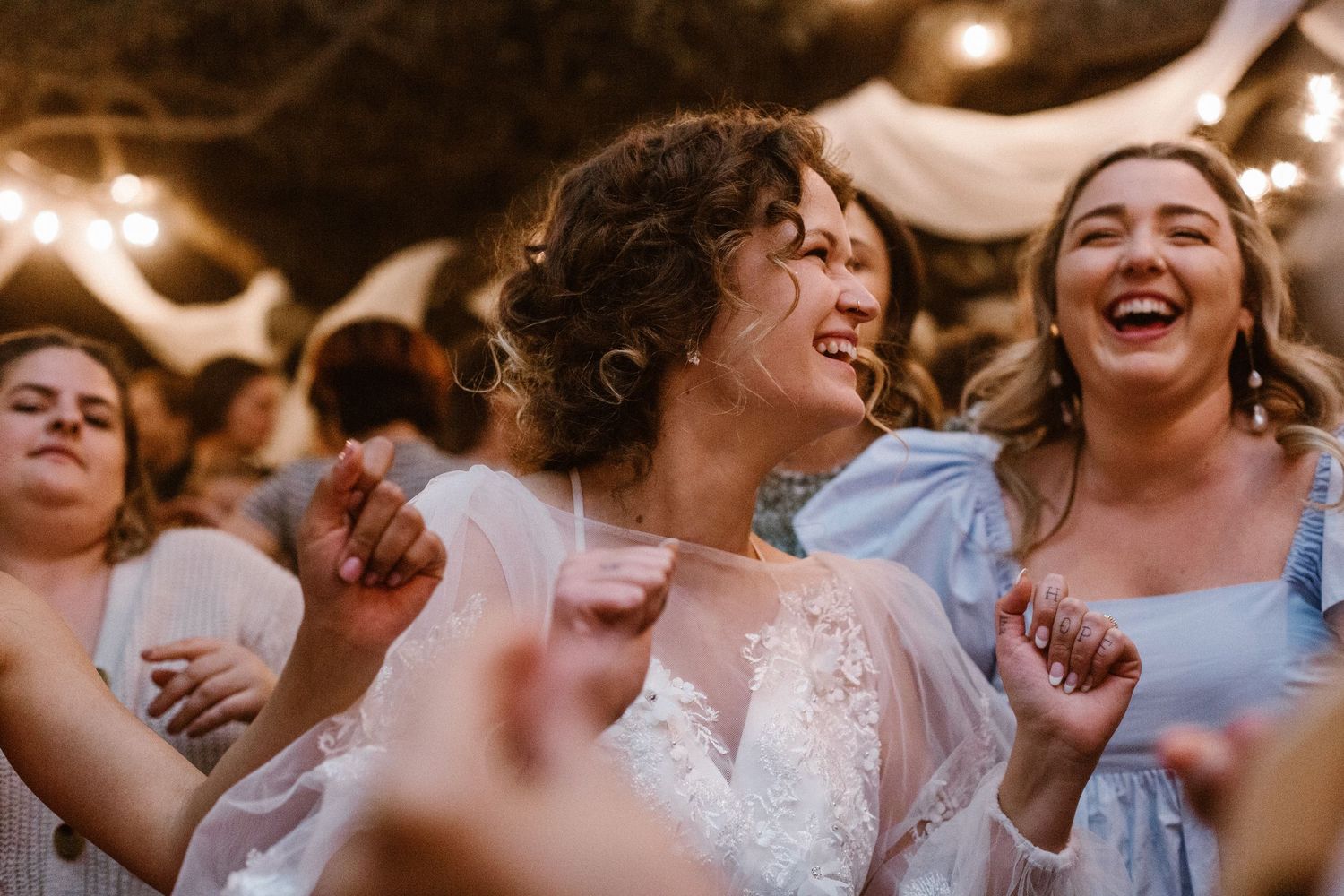 Bride in lace gown laughing and dancing with bridesmaids in blue at wedding reception.