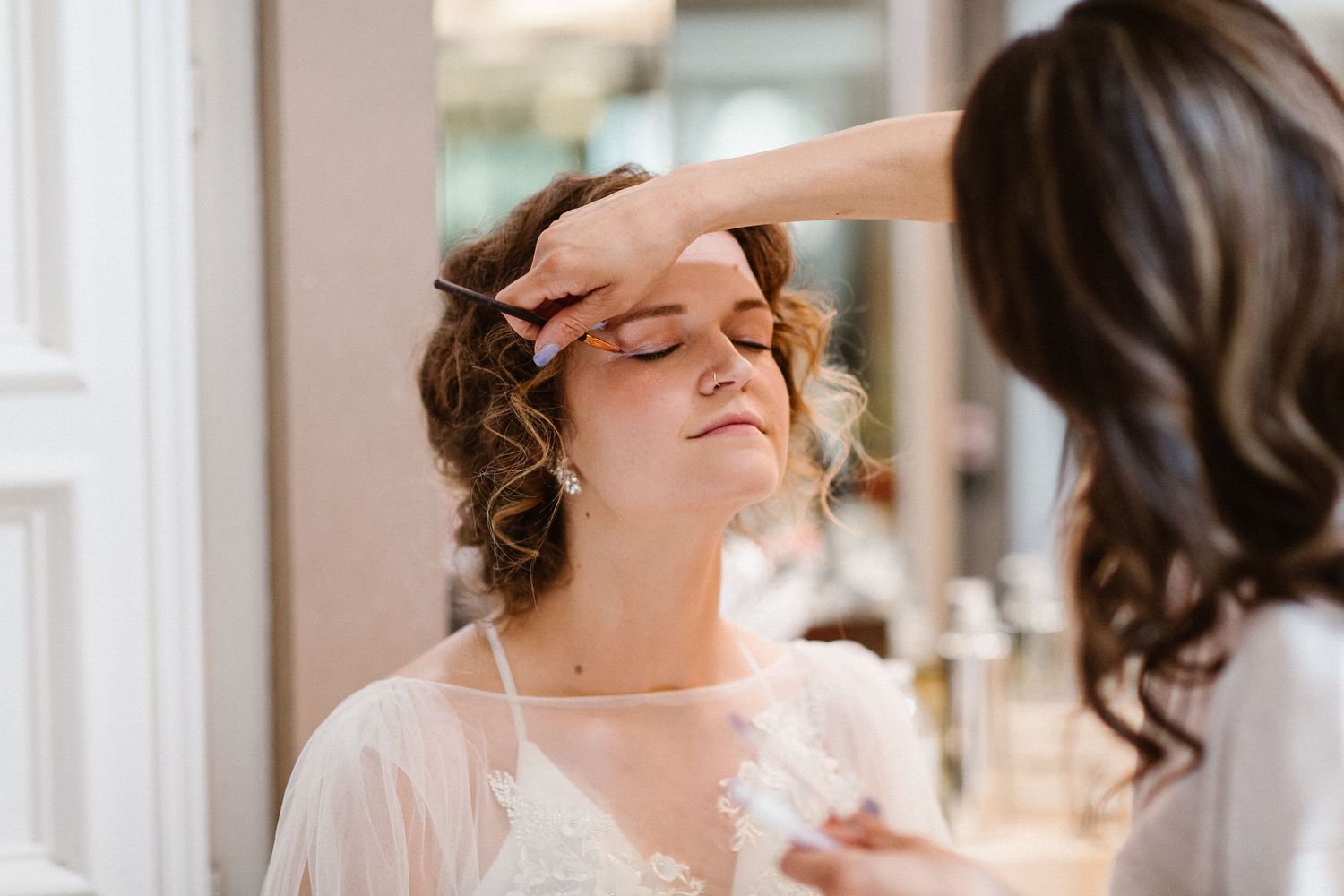 Makeup artist carefully applying bridal makeup to a bride in a white lace robe.
