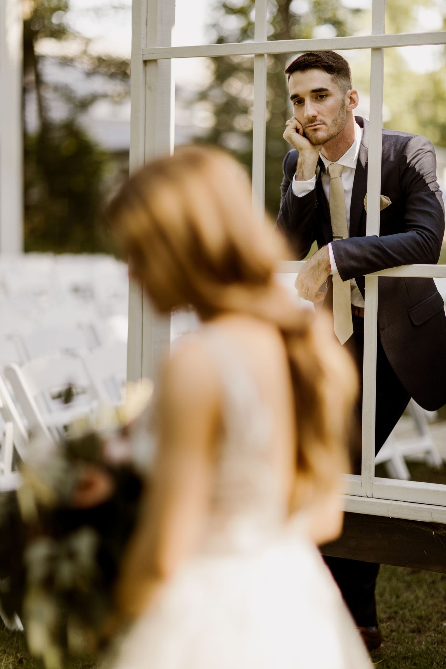 Groom in navy suit leans on white altar frame gazing at bride in foreground during outdoor ceremony.