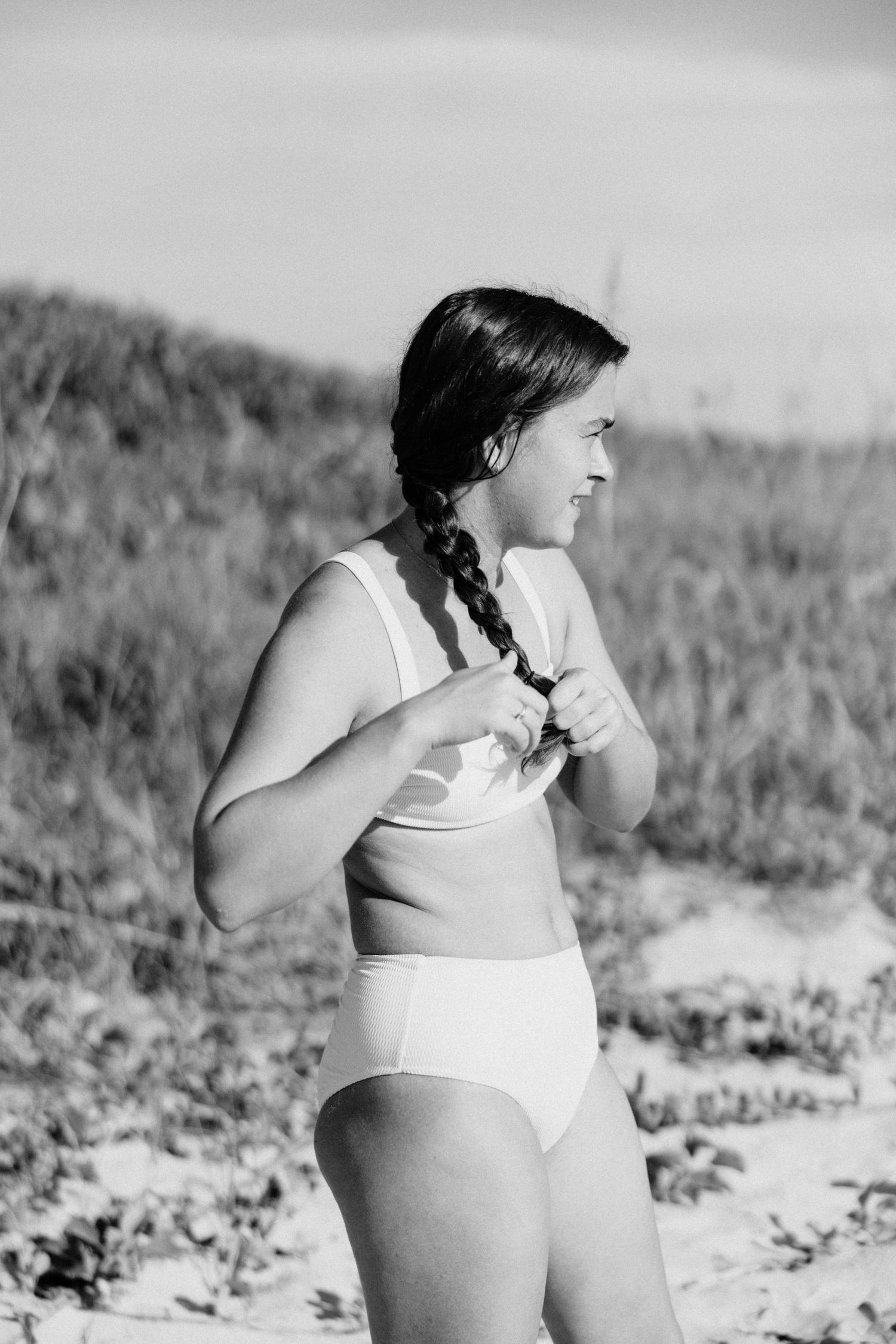 Black and white photo of woman in white swimsuit with braid standing in beach dunes.