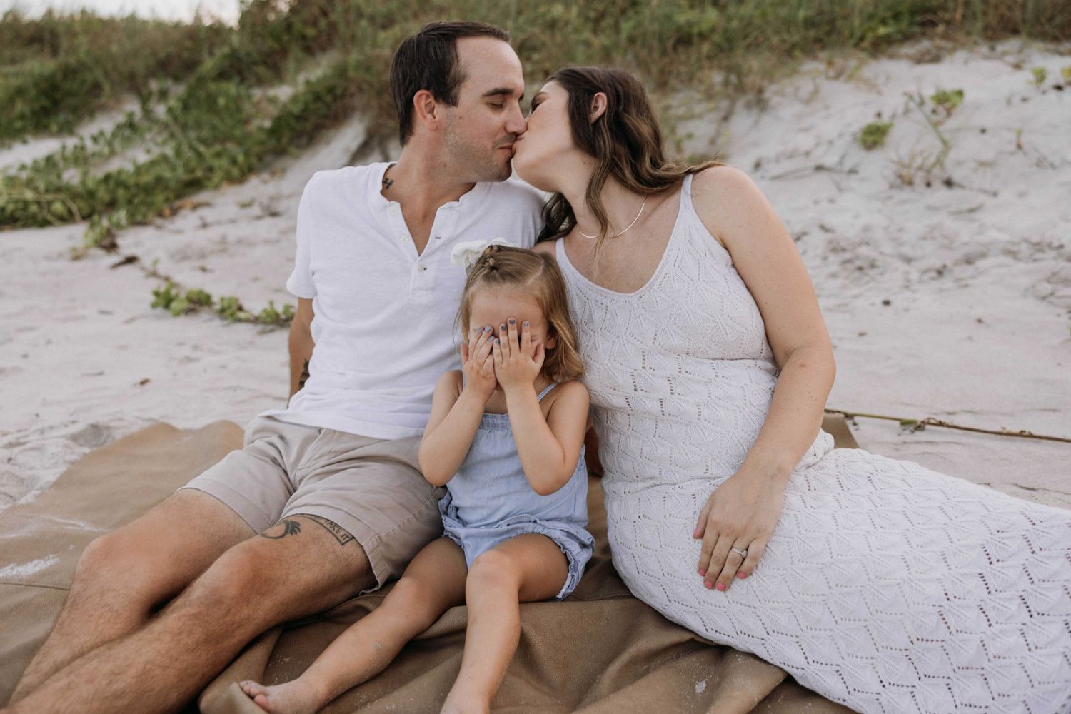 Couple kissing over their young daughter on a sandy beach during a warm family photo session.