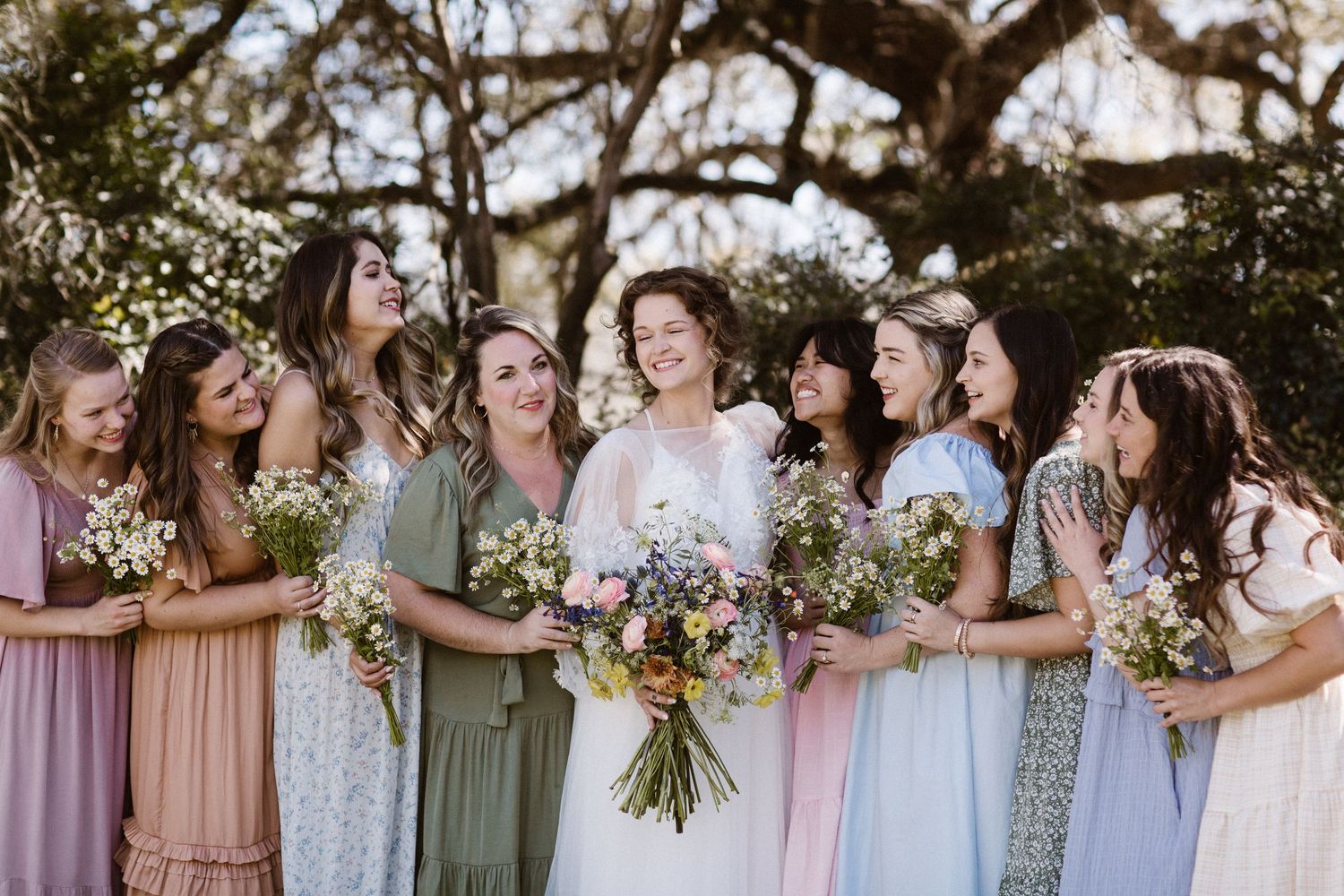 Smiling bride in white gown poses with eight bridesmaids in mismatched pastel dresses holding wildflower bouquets outdoors.