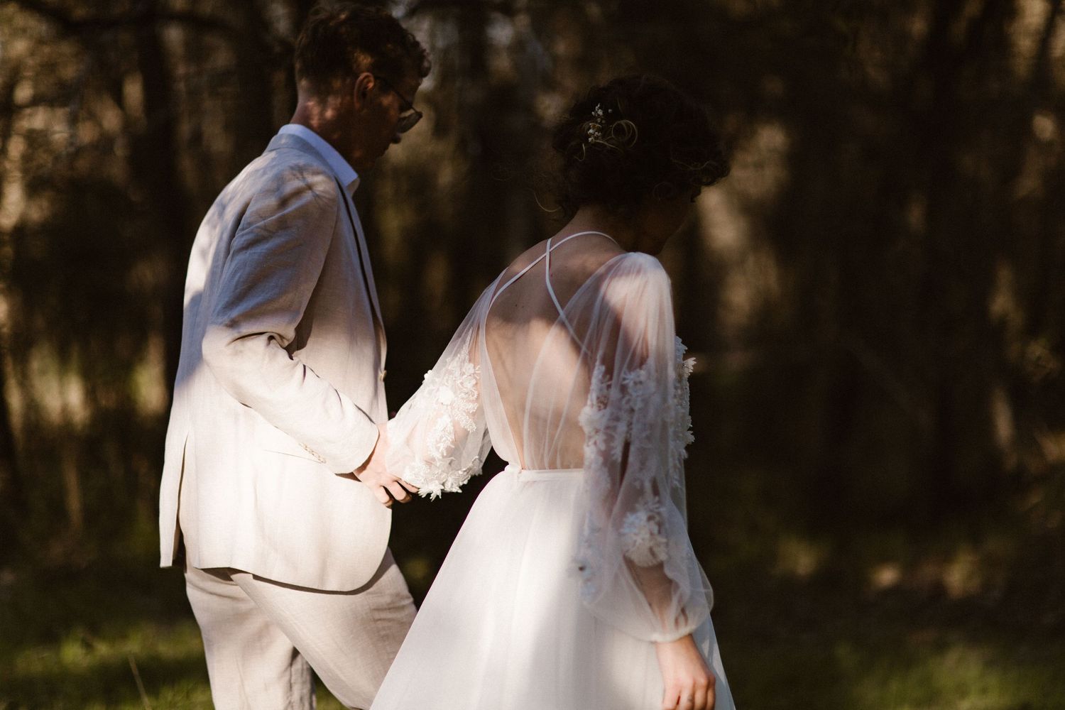Bride and groom holding hands walking through a sunlit forest on their wedding day.