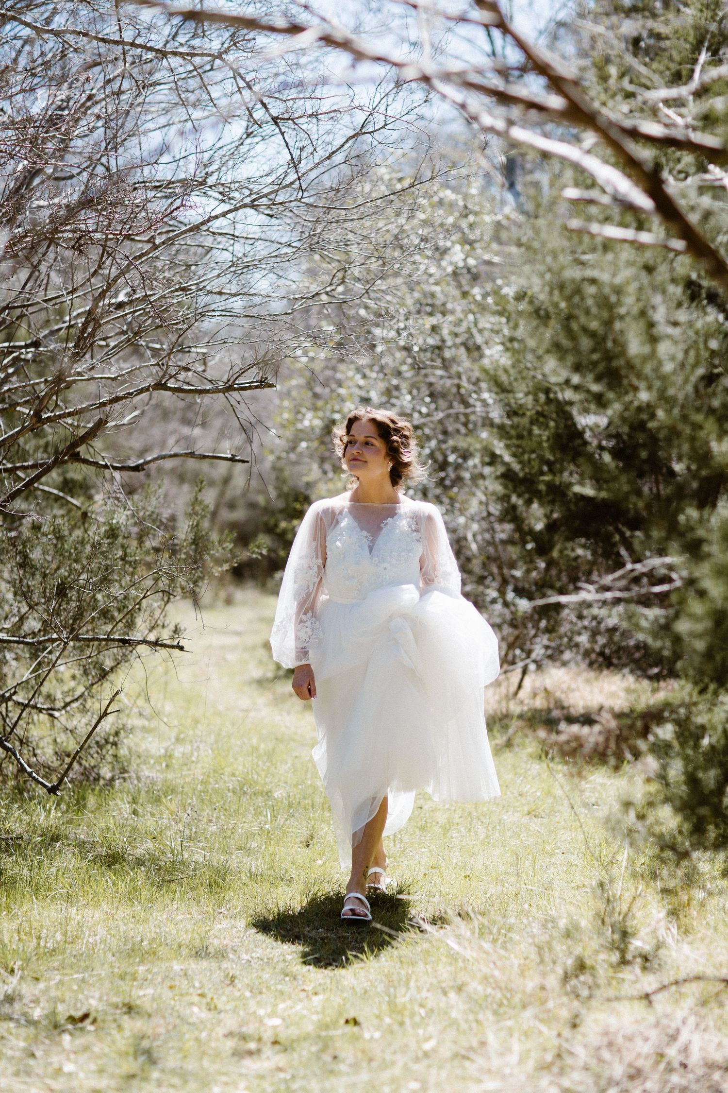 Bride in elegant white lace wedding dress walking through a sunlit outdoor garden setting.