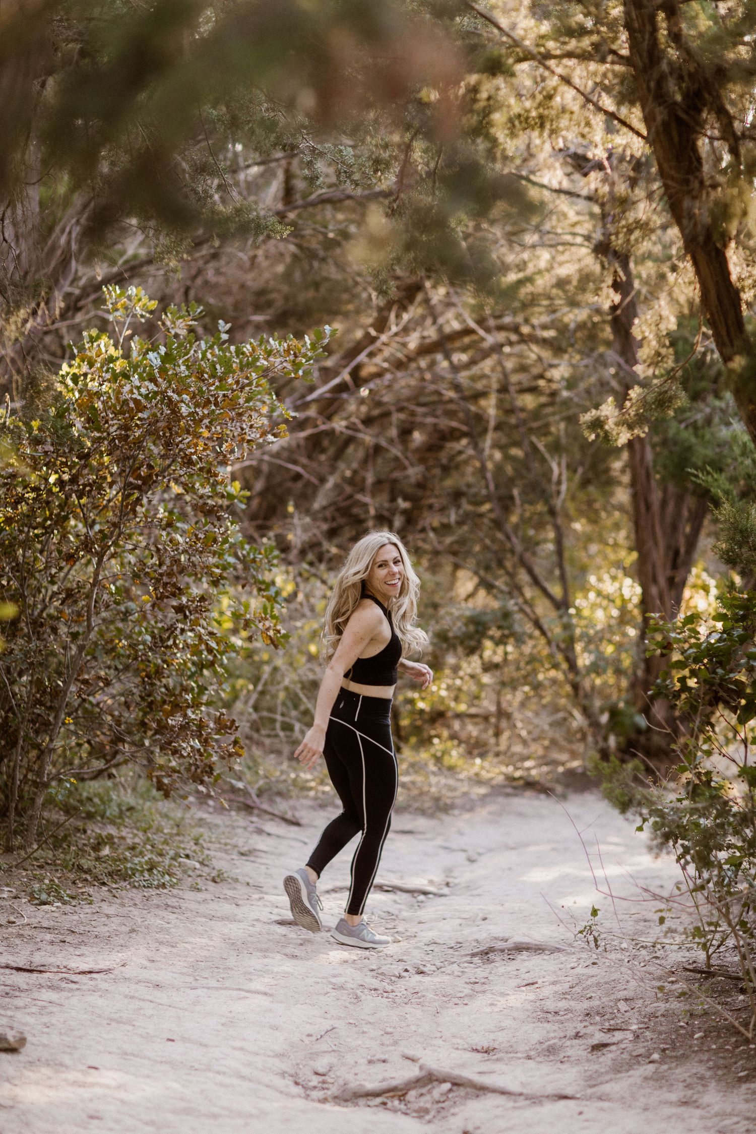 Blonde woman in black athletic wear walking on a sandy forest trail surrounded by lush green trees.