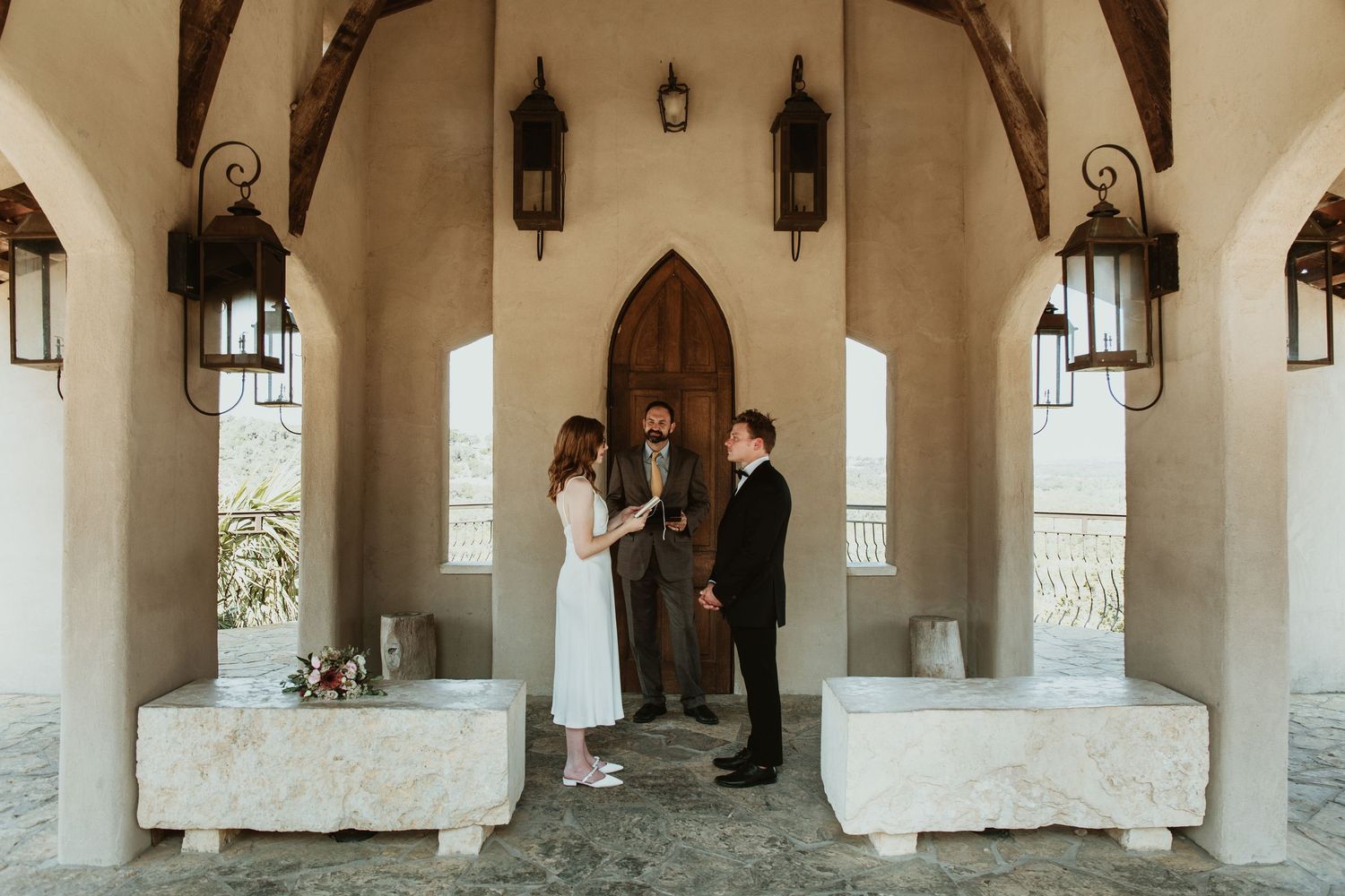 Couple exchanging vows under rustic arched pavilion with lanterns during intimate outdoor wedding ceremony.