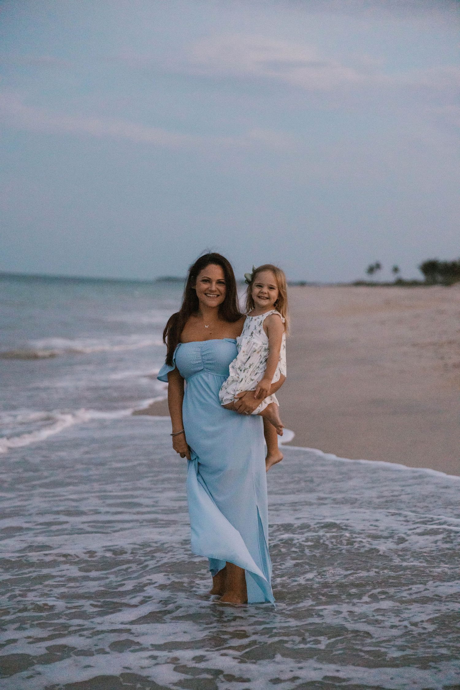 Woman in blue off-shoulder dress holds young girl on a beach at dusk.