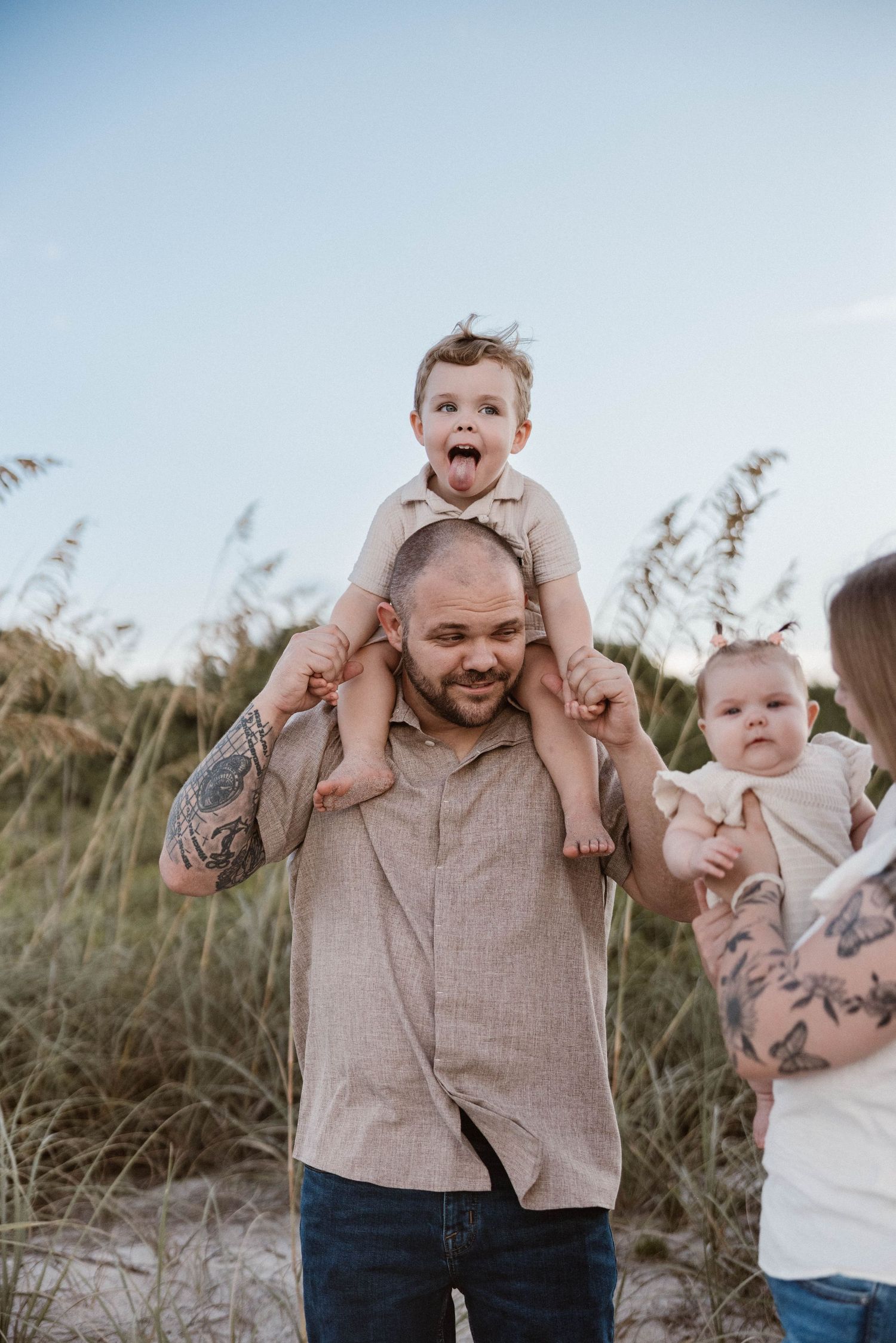Tattooed father with toddler on shoulders sticking tongue out, holding baby at sandy beach.