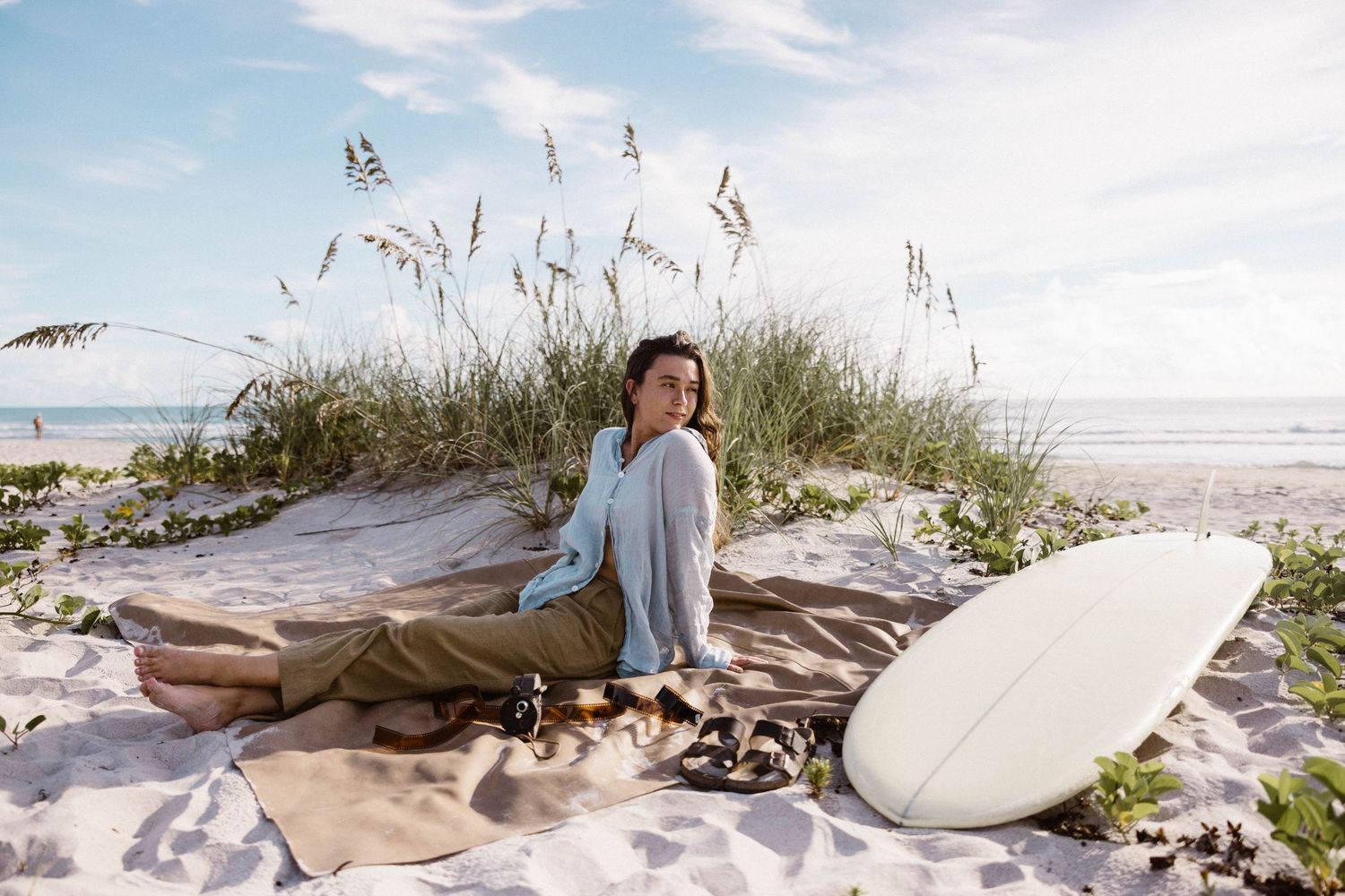 Young woman sitting on blanket at beach with surfboard and sea oats in background.