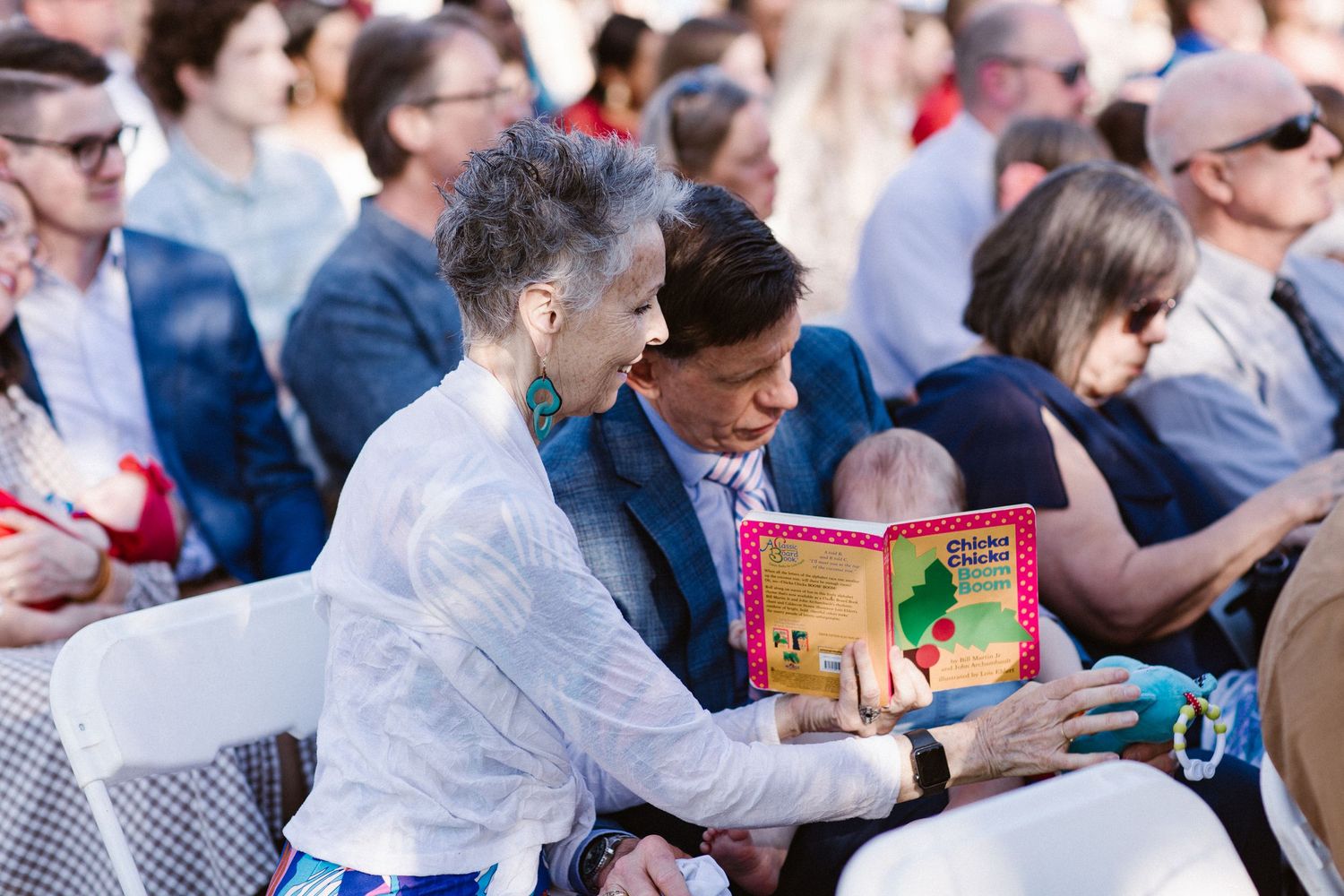 Two women share a children's book while seated among guests at an outdoor wedding ceremony.
