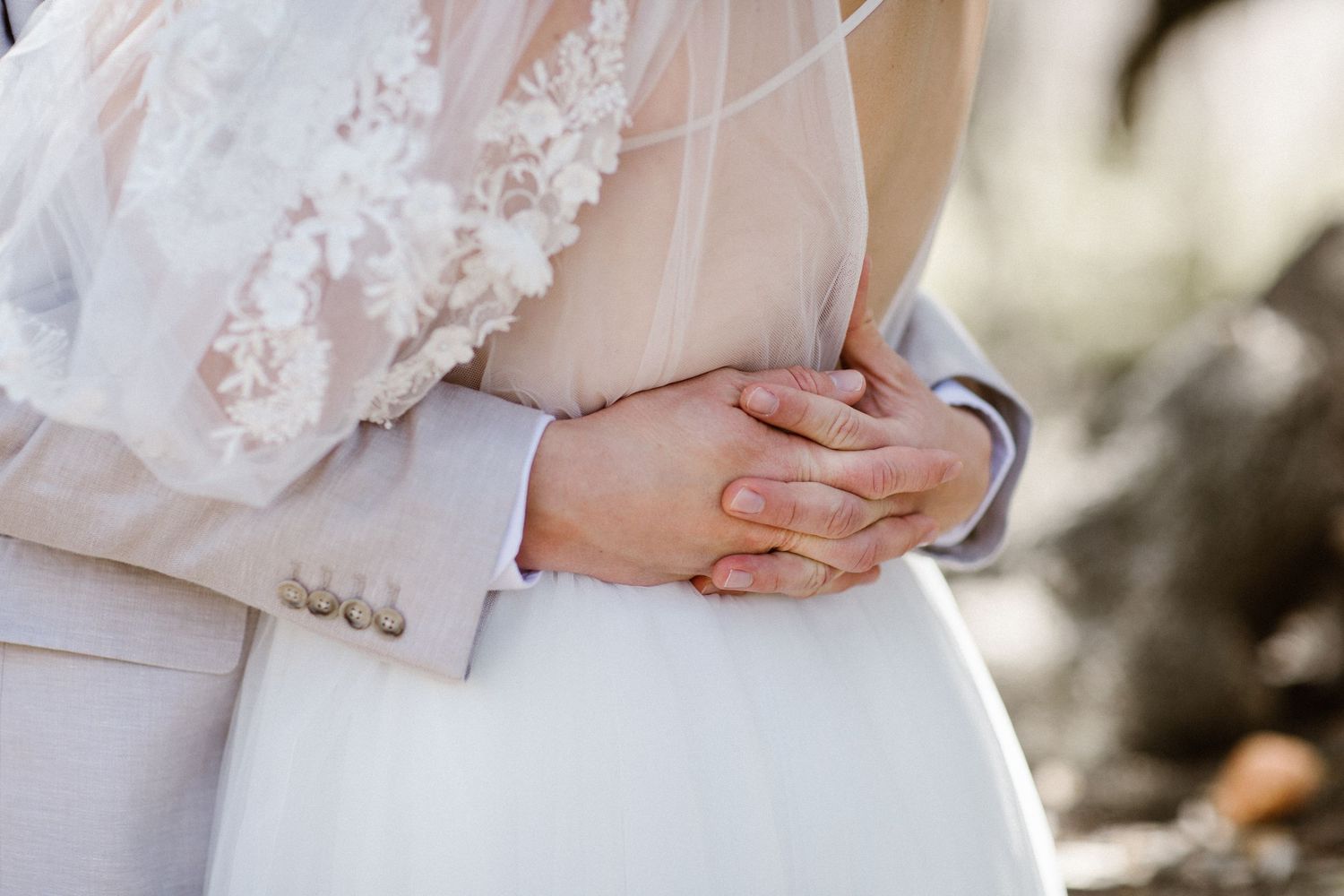 Close-up of groom gently holding bride around waist, hands clasped over white wedding gown.