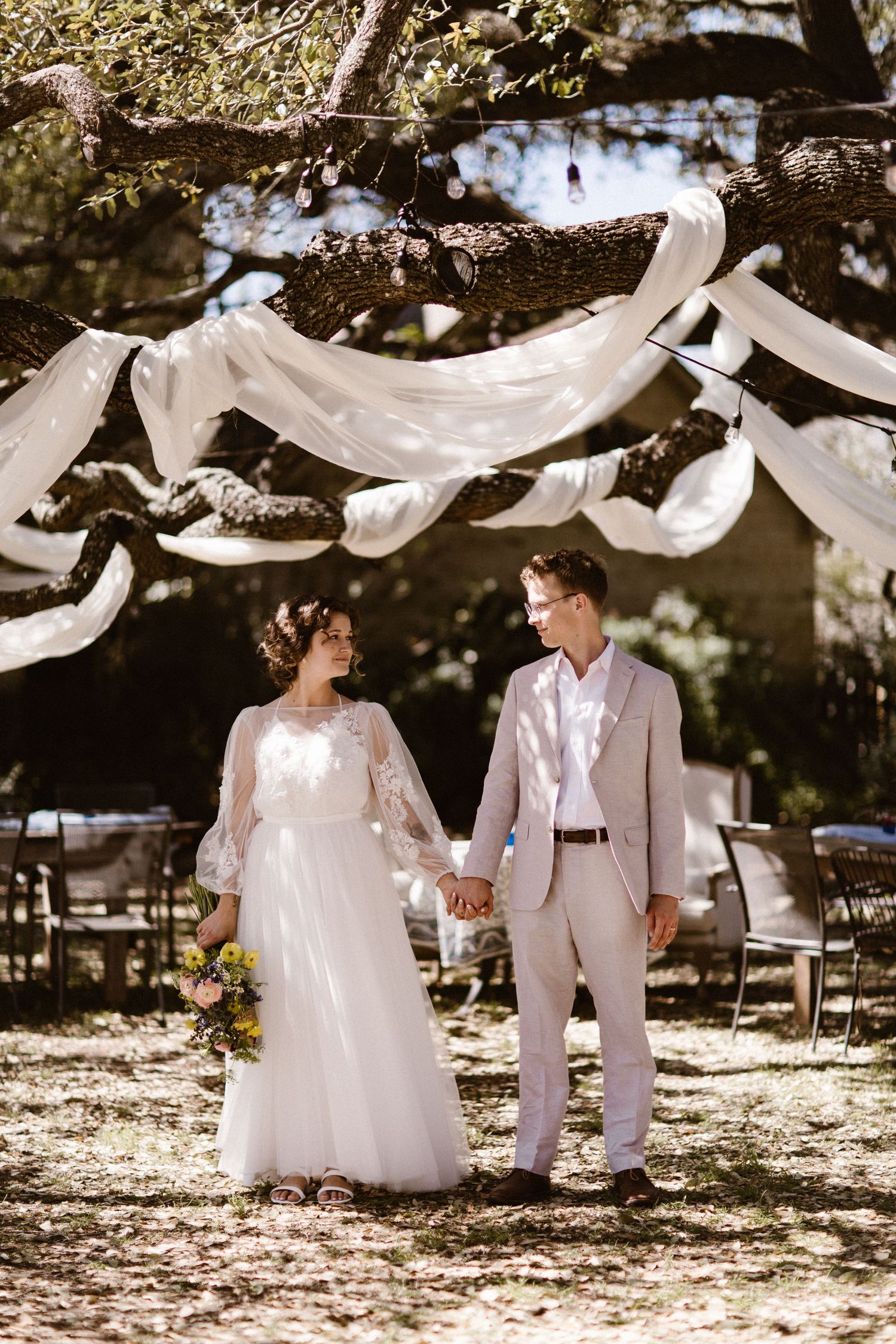Bride and groom holding hands under white draped tree at outdoor wedding ceremony.