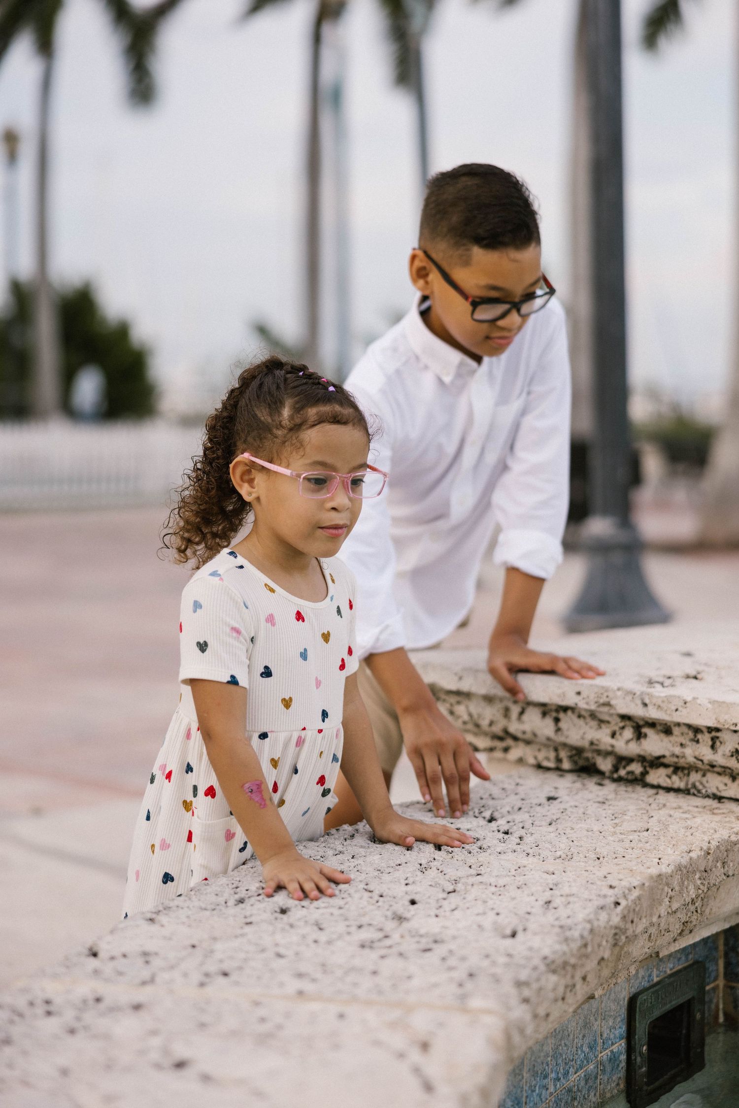 Two children with glasses explore a stone ledge outdoors near palm trees.