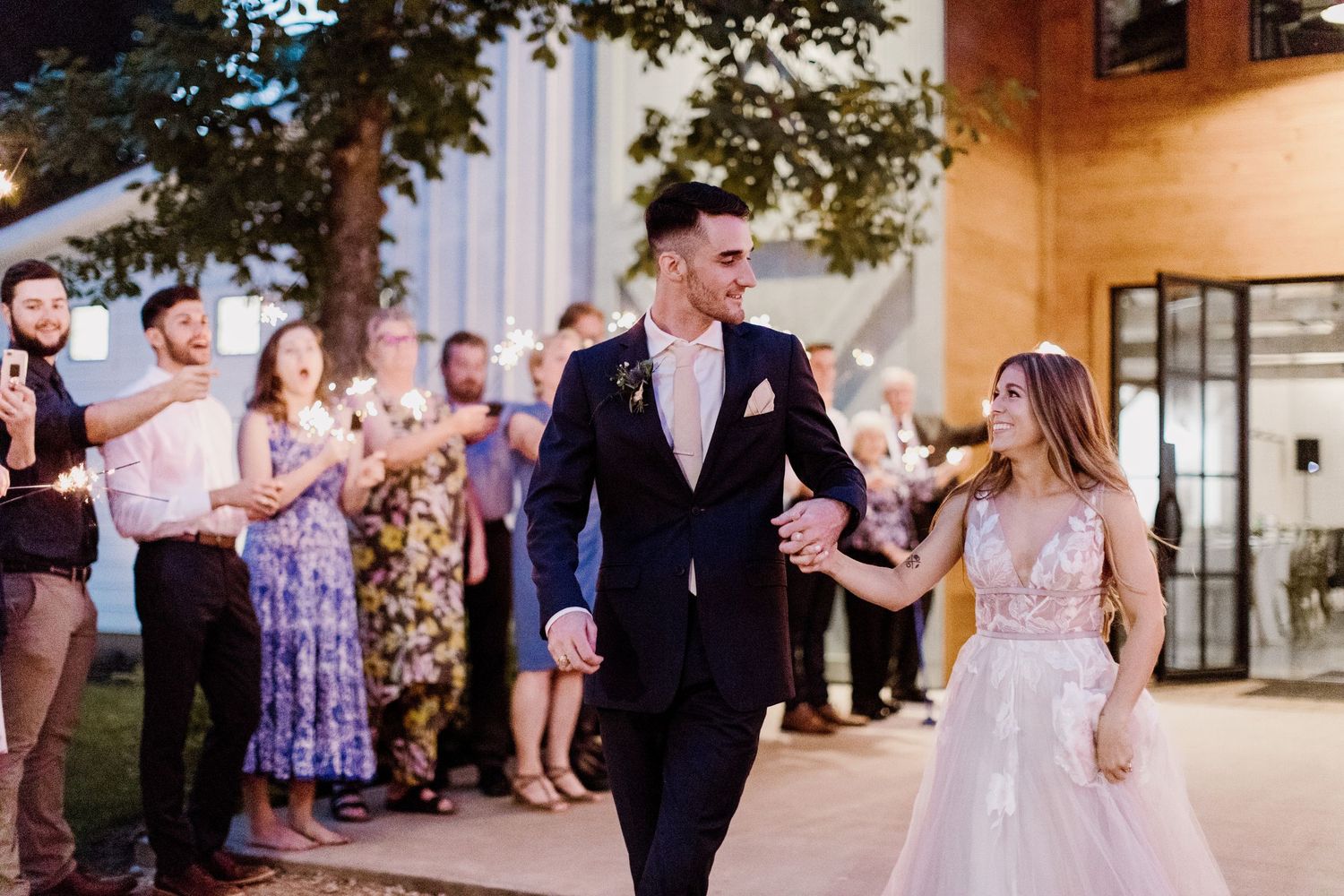 Newlyweds walk hand-in-hand through sparkler send-off surrounded by cheering guests at elegant evening wedding reception.
