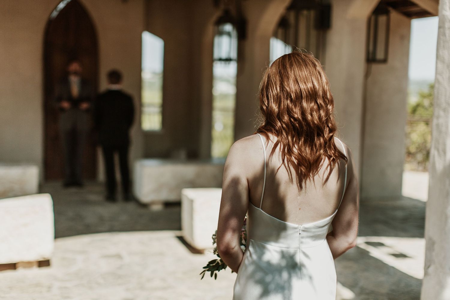 Bride in white dress with red hair holds bouquet, facing groom at outdoor wedding chapel entrance.