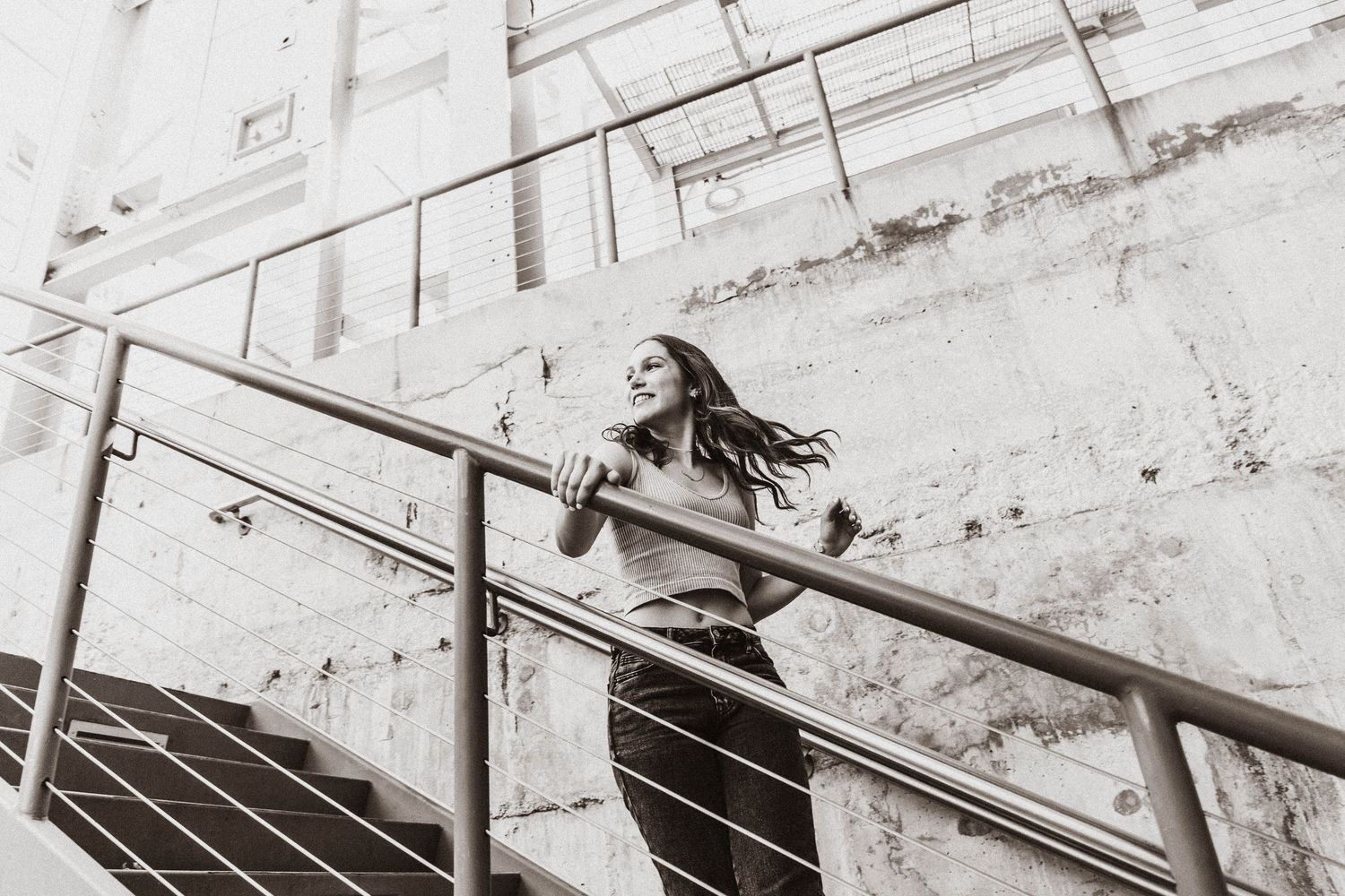 Black and white photo of a young woman with flowing hair standing on a staircase railing outdoors.