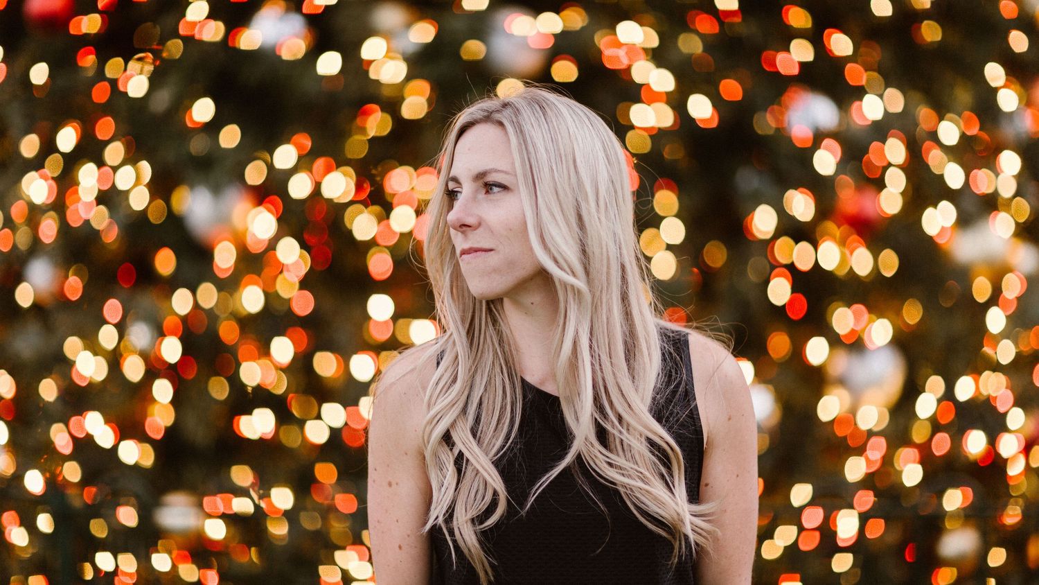 Blonde woman in black dress posing against a festive bokeh Christmas lights background.