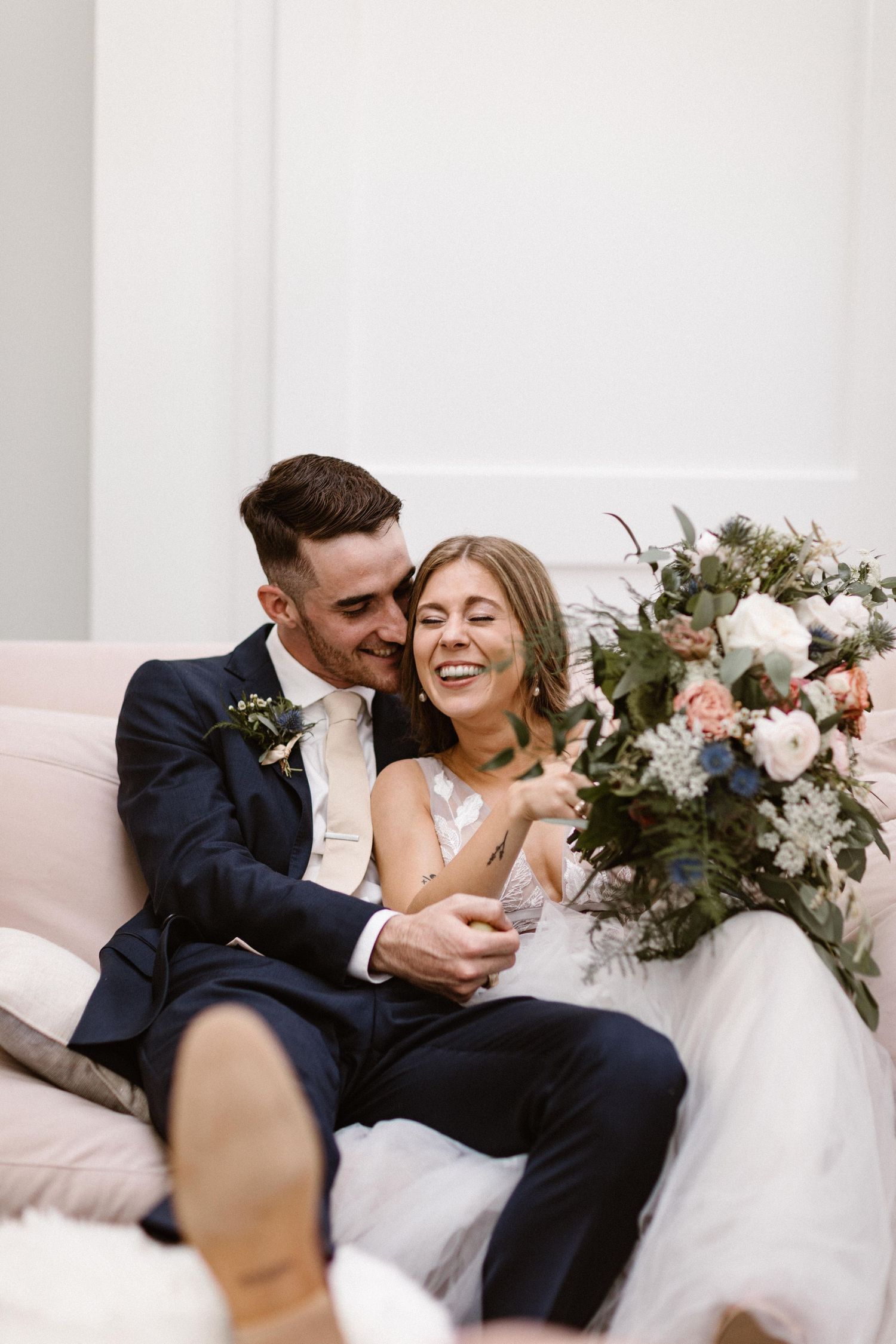 Laughing bride and groom sitting together on a pink sofa with floral bouquet.