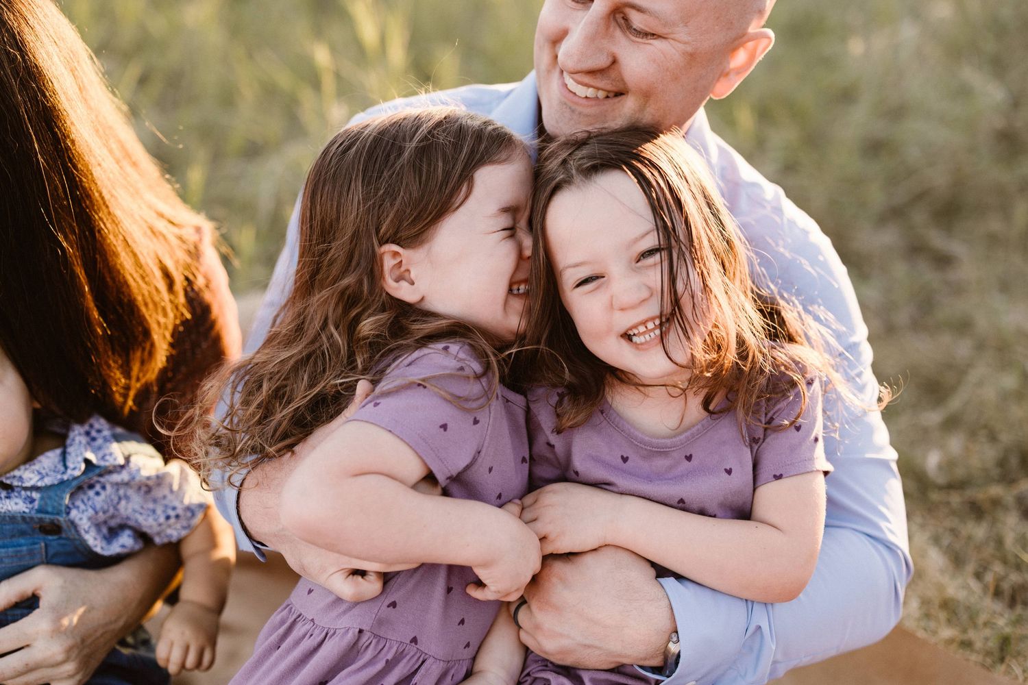 Father sitting outdoors hugging two smiling daughters in matching purple dresses in golden light.