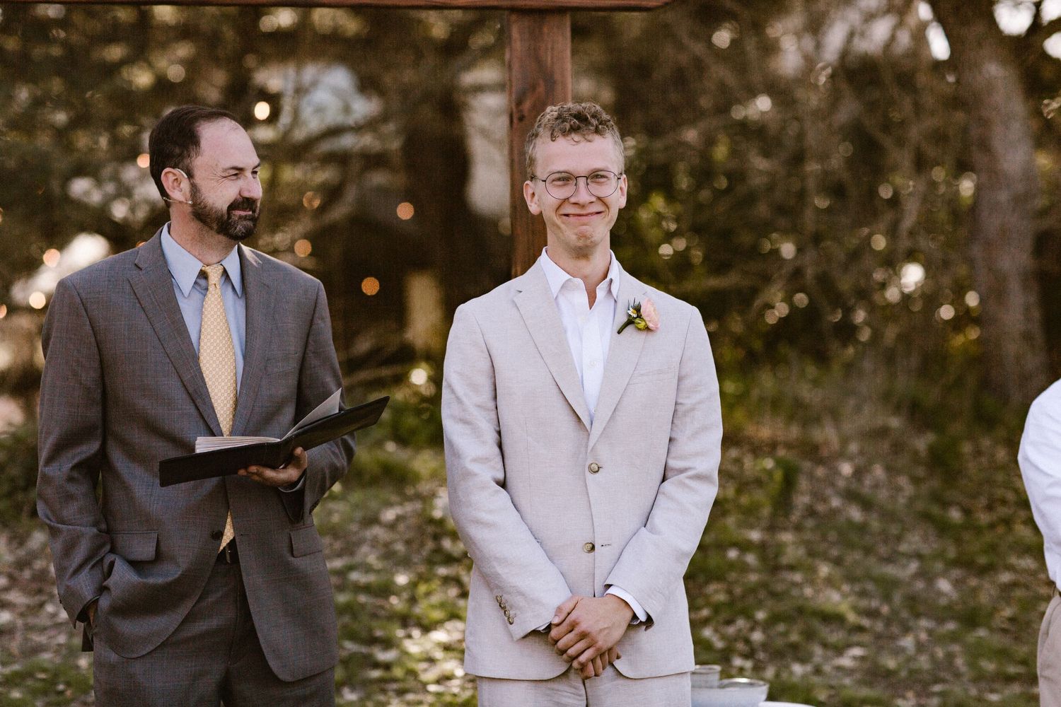 Groom in a light gray suit smiles with clasped hands as he waits at the altar during an outdoor wedding ceremony.