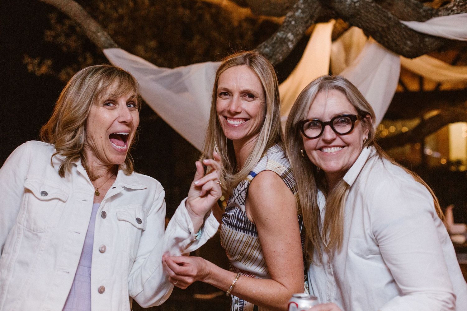 Three women in white jackets smiling and posing together at an outdoor evening event.
