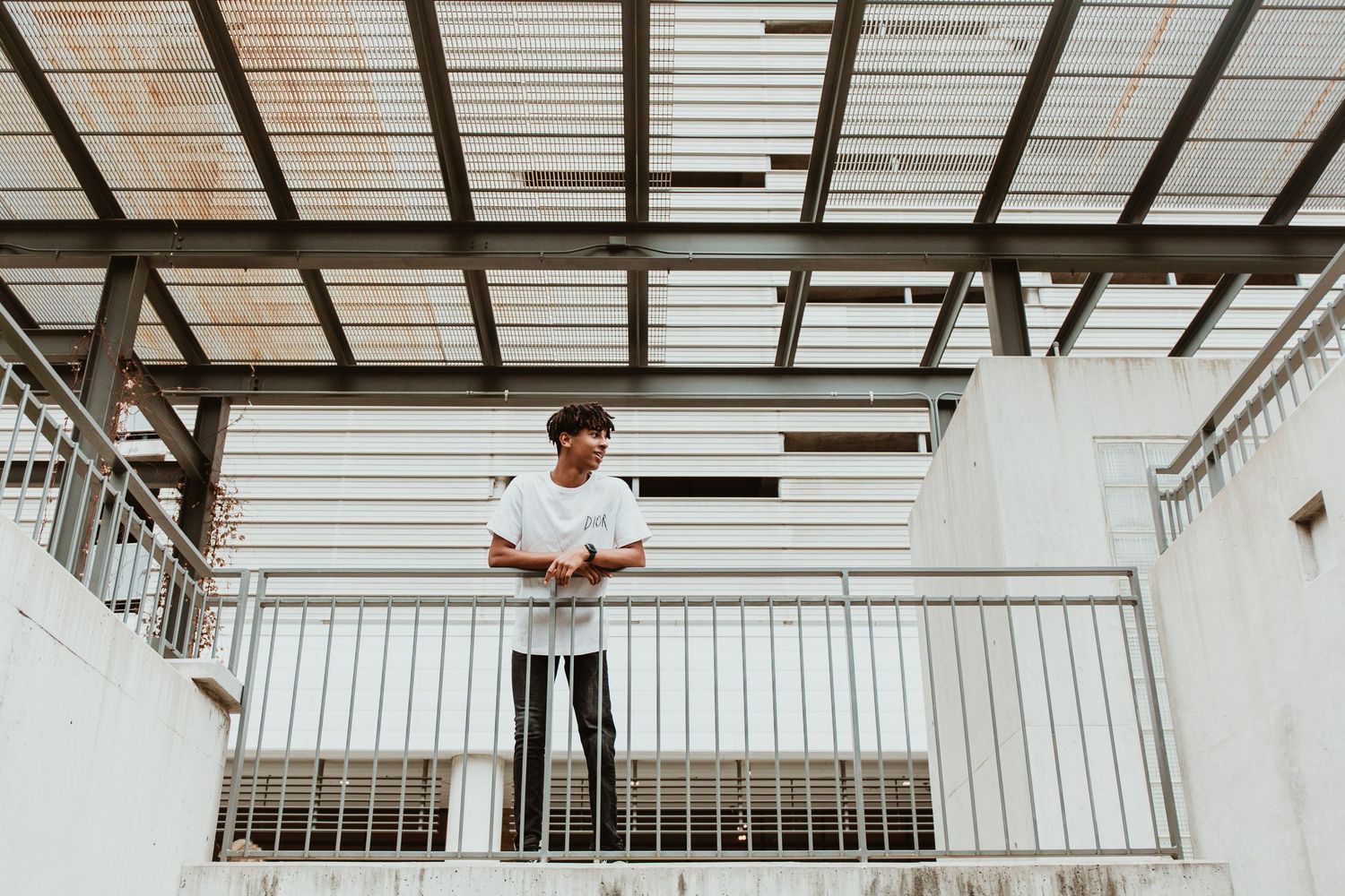 Young man in white t-shirt leaning on metal railing on elevated platform with industrial ceiling.