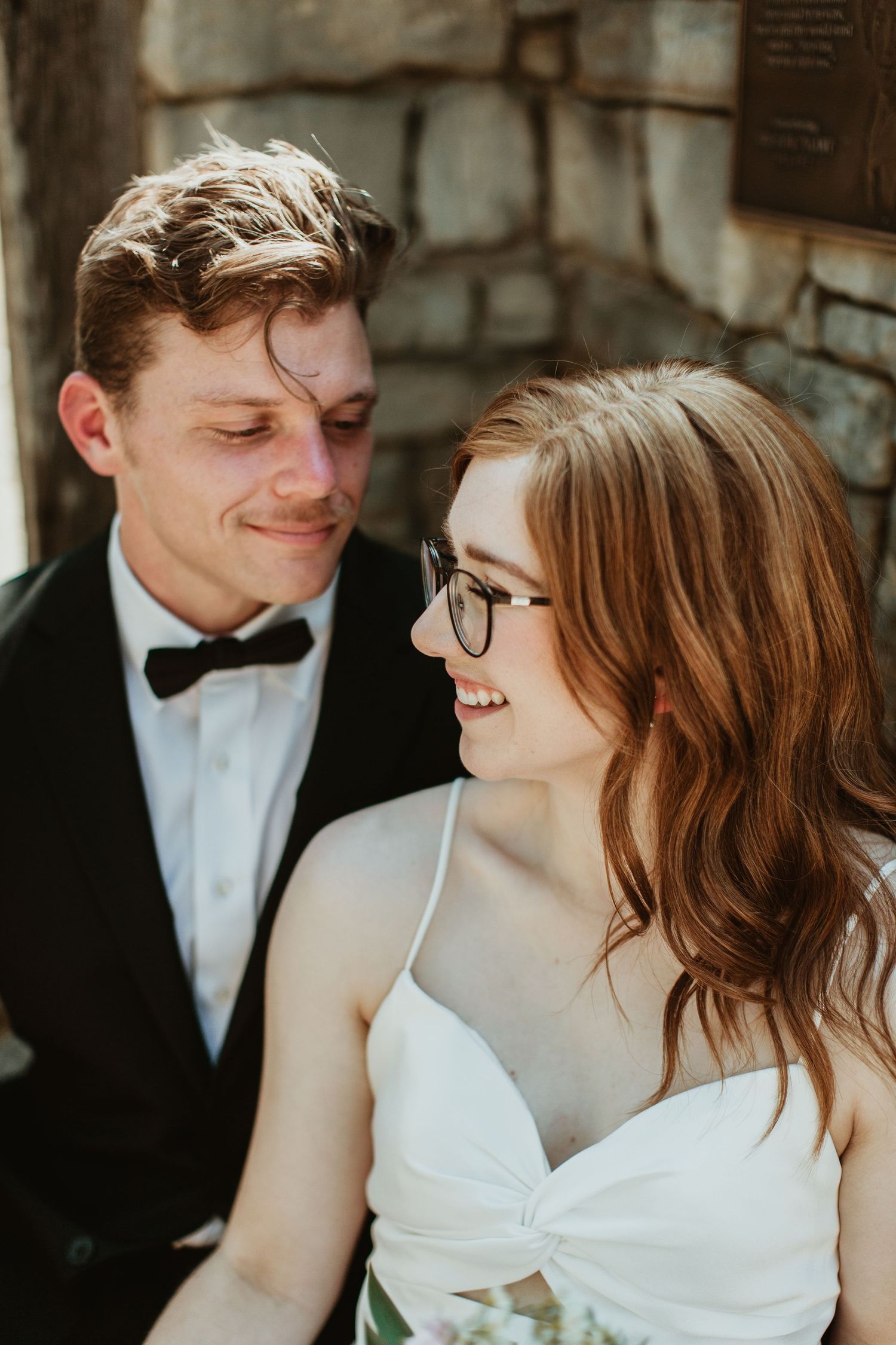A smiling bride in a white dress and glasses gazes at her groom in a black tuxedo and bow tie outdoors.