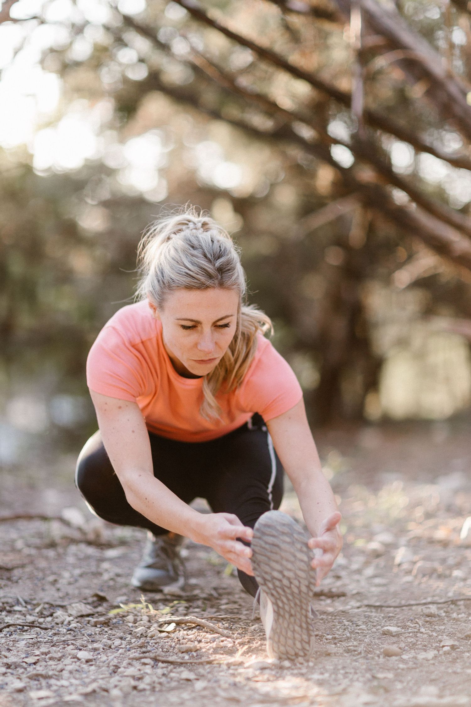 Woman in pink top stretching before a trail run in a sunlit forest setting.