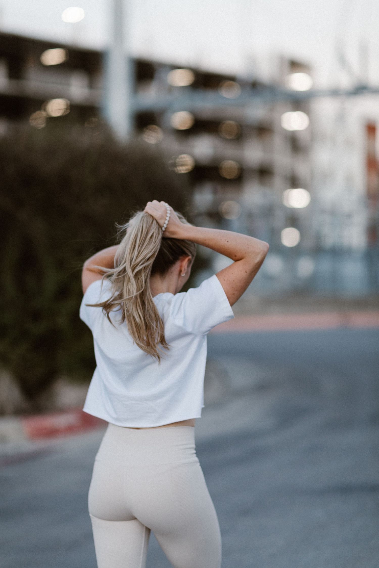 Woman in white crop top and beige leggings fixing her blonde ponytail outdoors near urban building.
