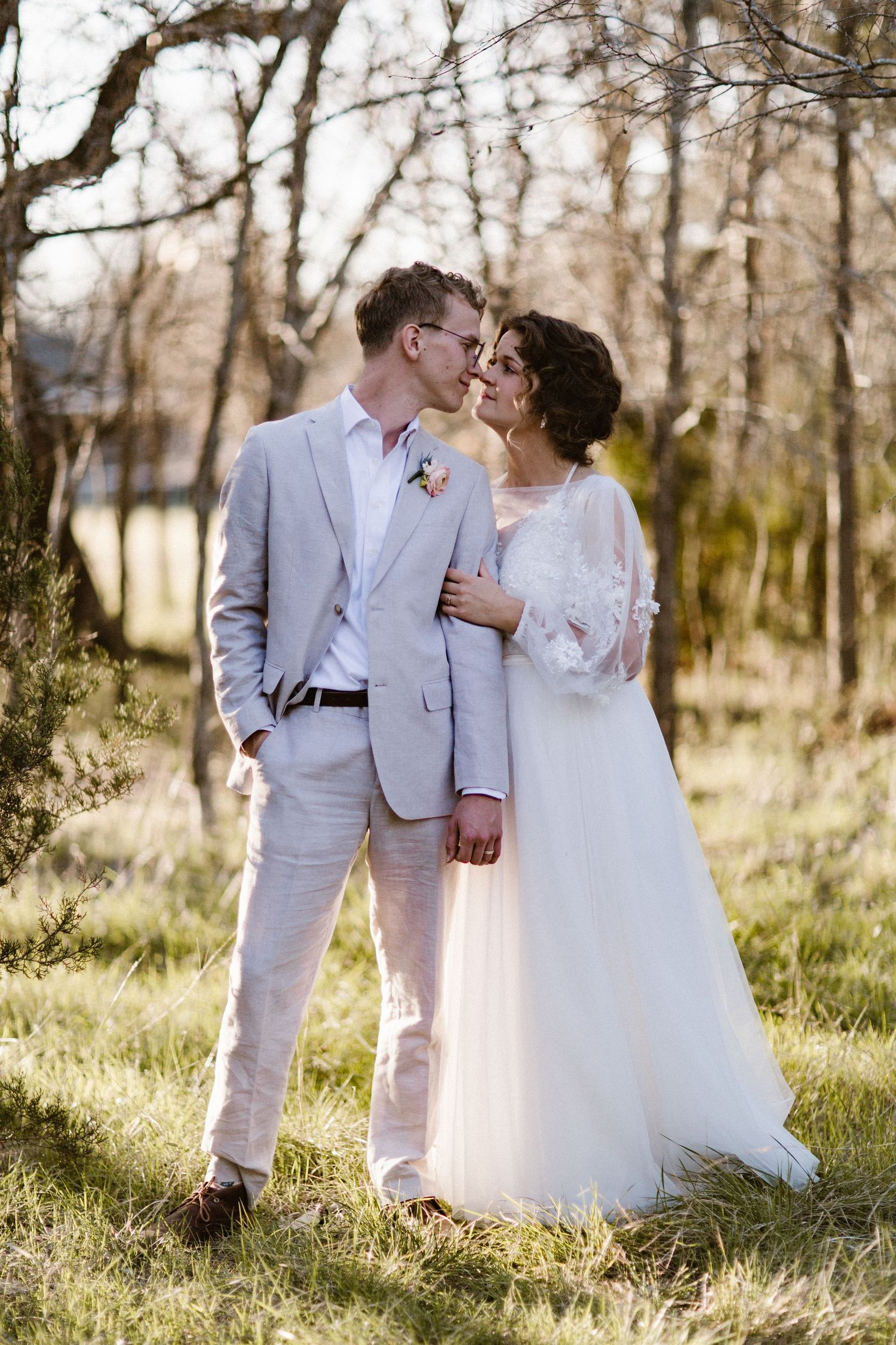 Bride and groom sharing a romantic kiss in sunlit outdoor setting, groom in gray suit and bride in white dress.