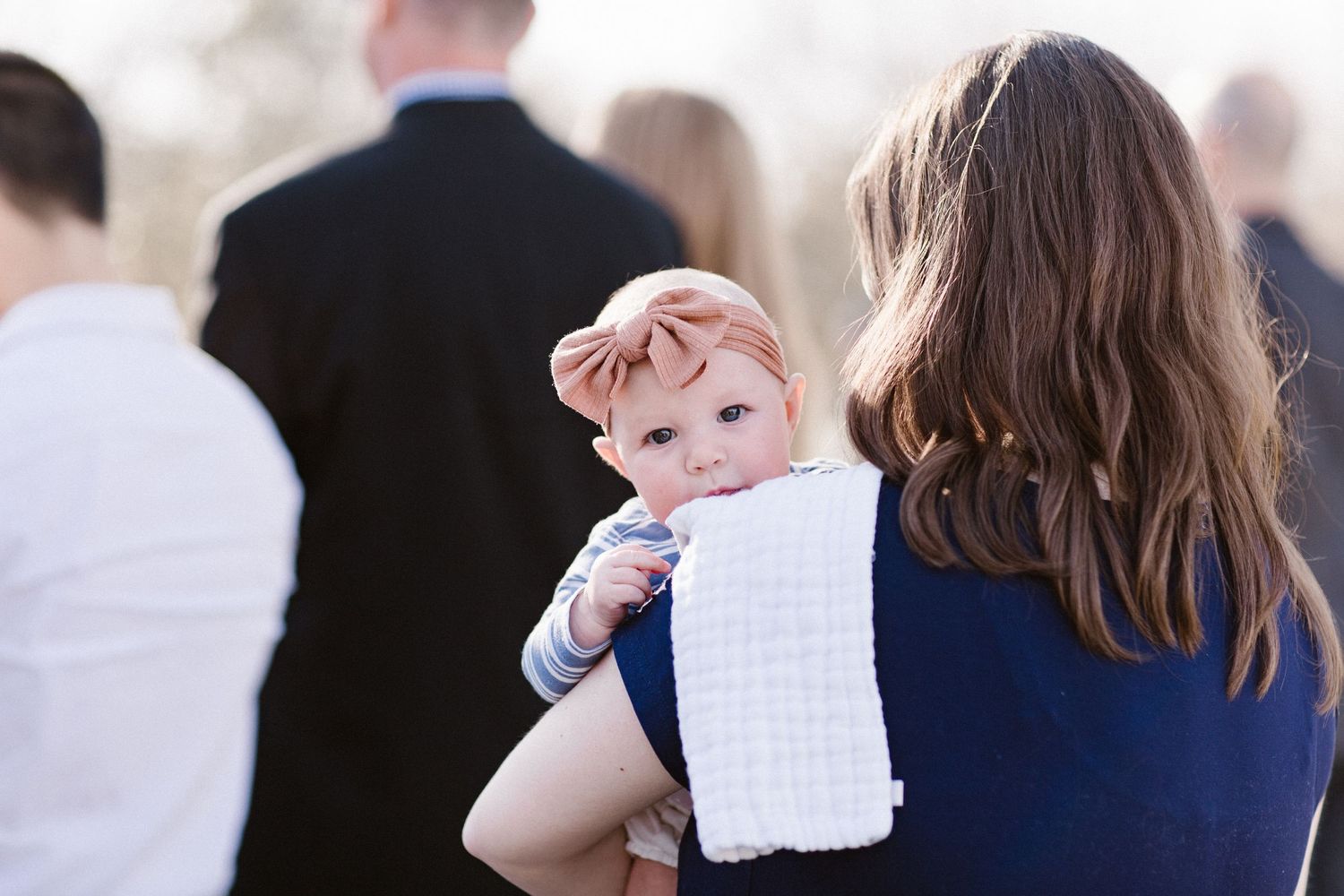 Adorable red-haired baby in blue outfit resting on mothers shoulder during outdoor wedding ceremony.