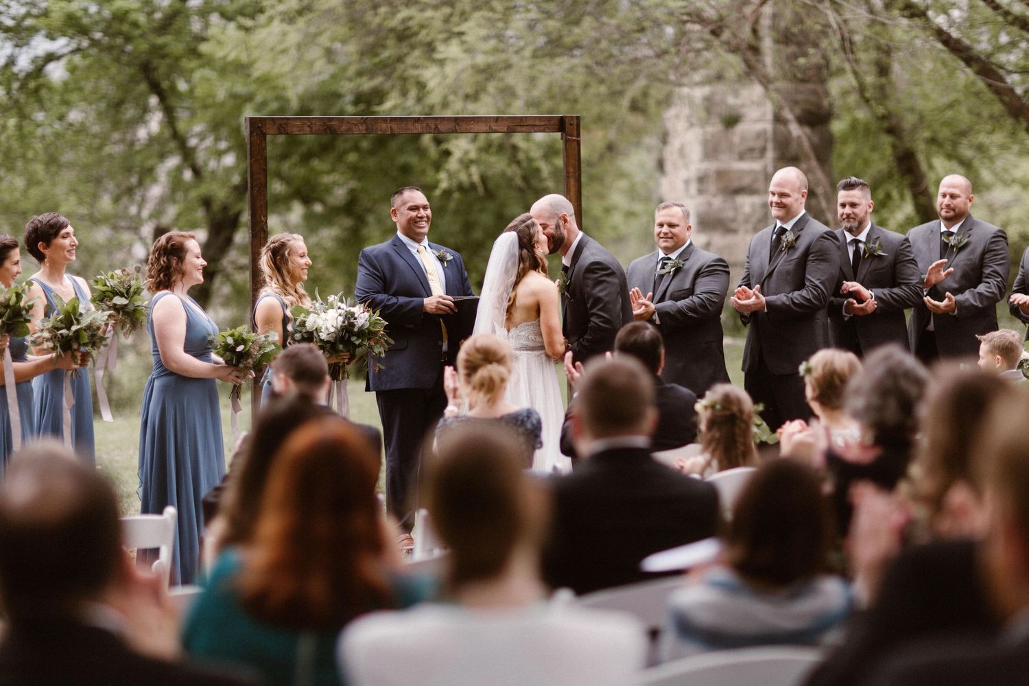 A bride and groom share a first kiss at an outdoor wedding ceremony with guests.