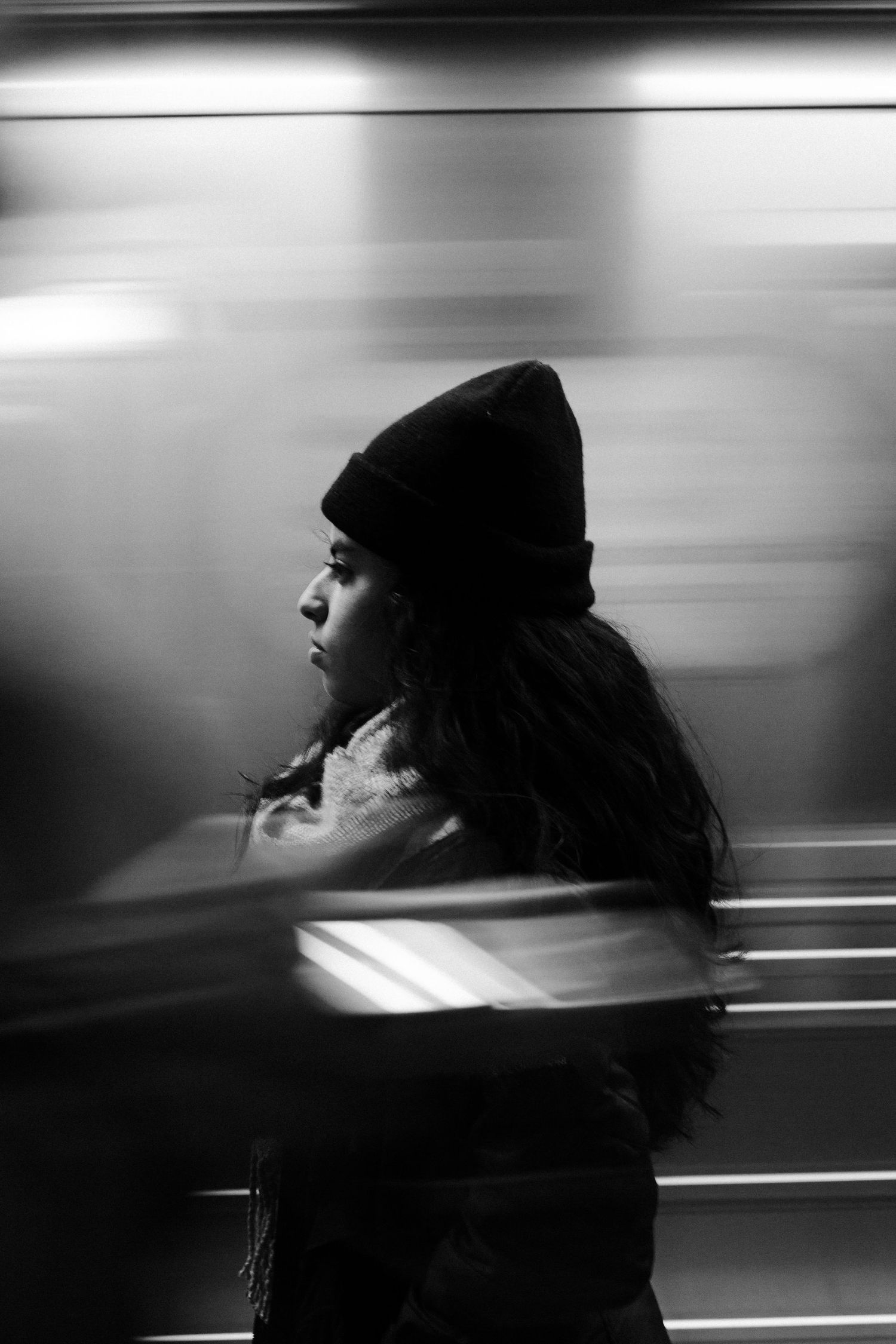 Black and white portrait of woman in beanie hat with motion-blurred subway train.