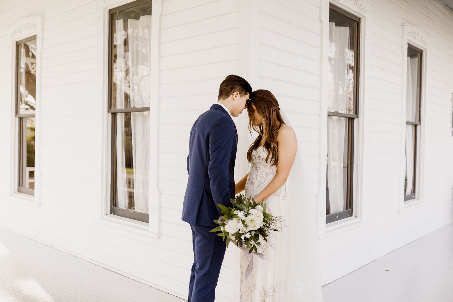 Bride and groom sharing intimate moment outside white farmhouse building, bride holding white bouquet.