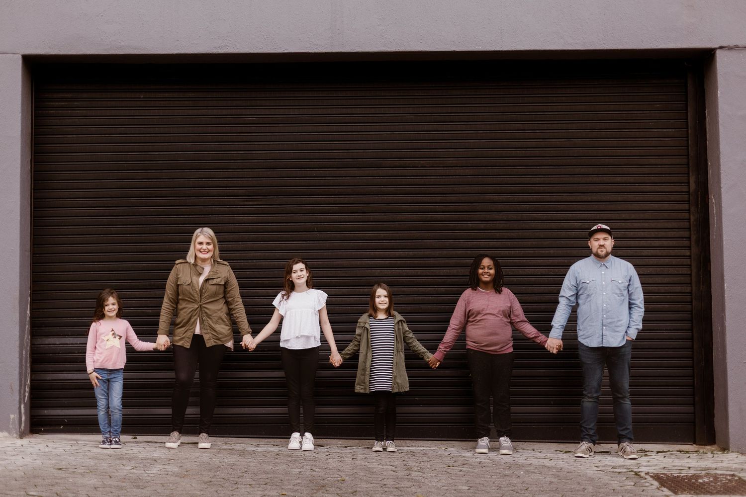 Blended family of six holding hands in a line in front of a dark industrial garage door.