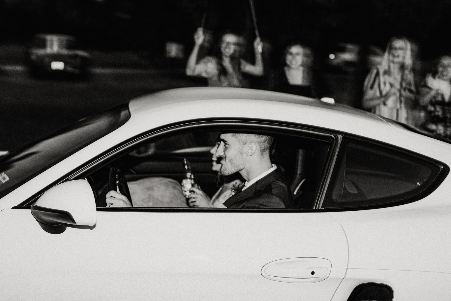 Black and white photo of a groom sitting in a white sports car holding drinks while guests celebrate in the background.