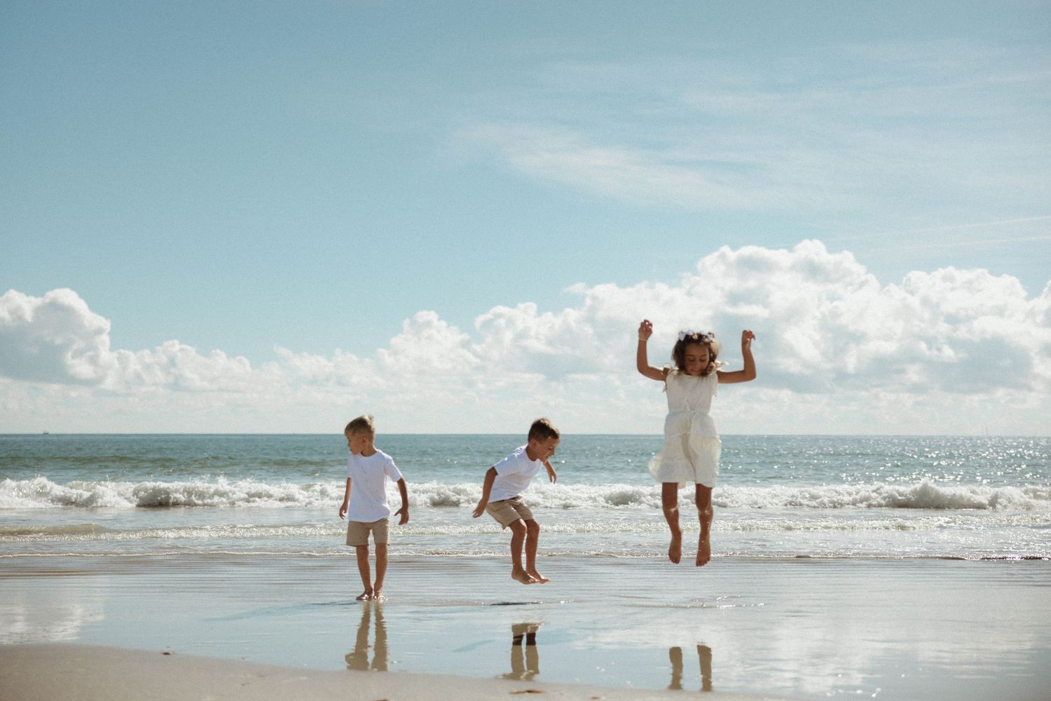 Three children jumping and playing joyfully on a sunny beach with ocean waves in the background.