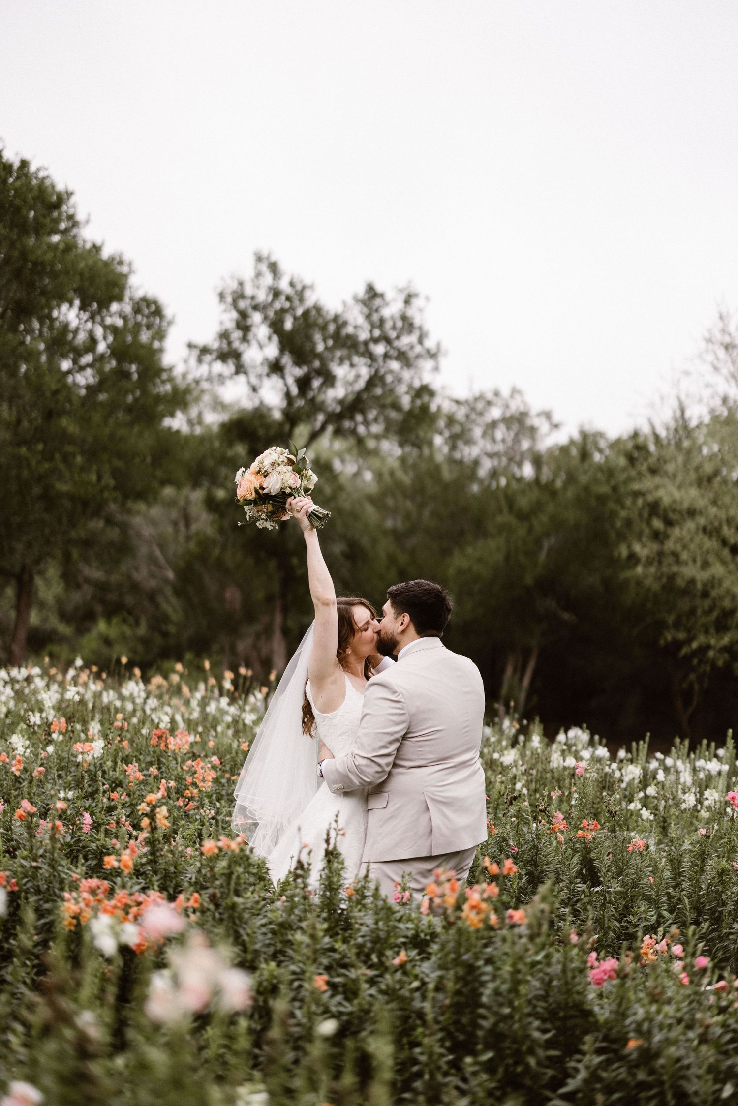 Bride raises bouquet while kissing groom in colorful wildflower field during outdoor wedding.