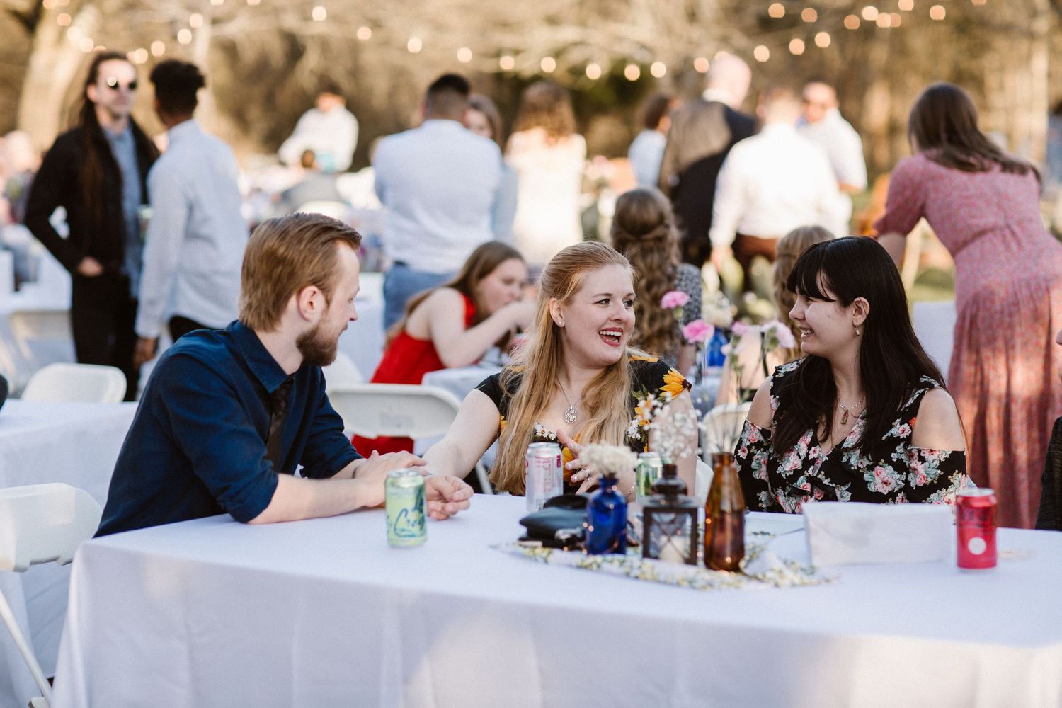Three guests laugh and chat together at a decorated table during an outdoor wedding reception.
