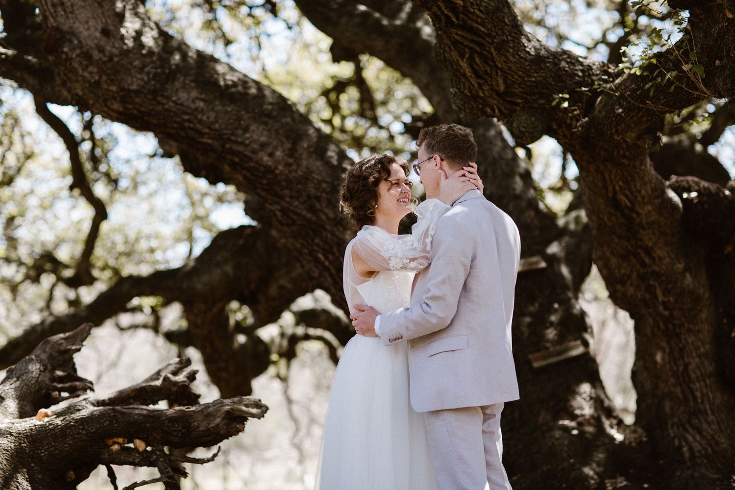 Bride and groom embrace romantically beneath sprawling oak tree branches in outdoor wedding portrait.