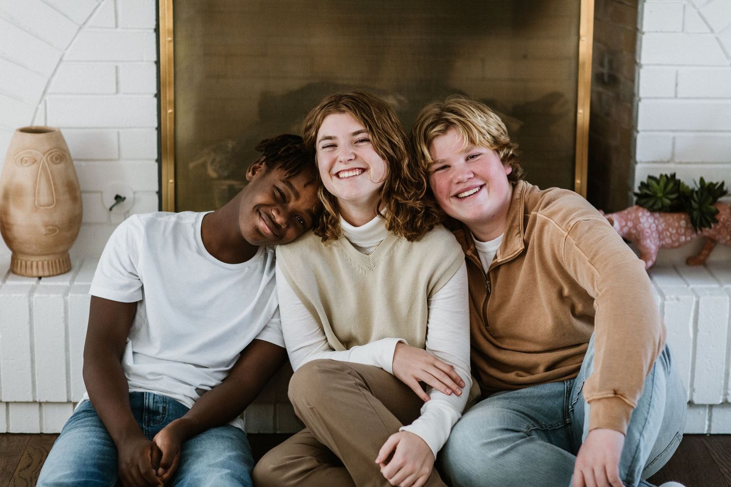 Three smiling teens sit together in front of a fireplace, sharing a joyful moment of friendship.